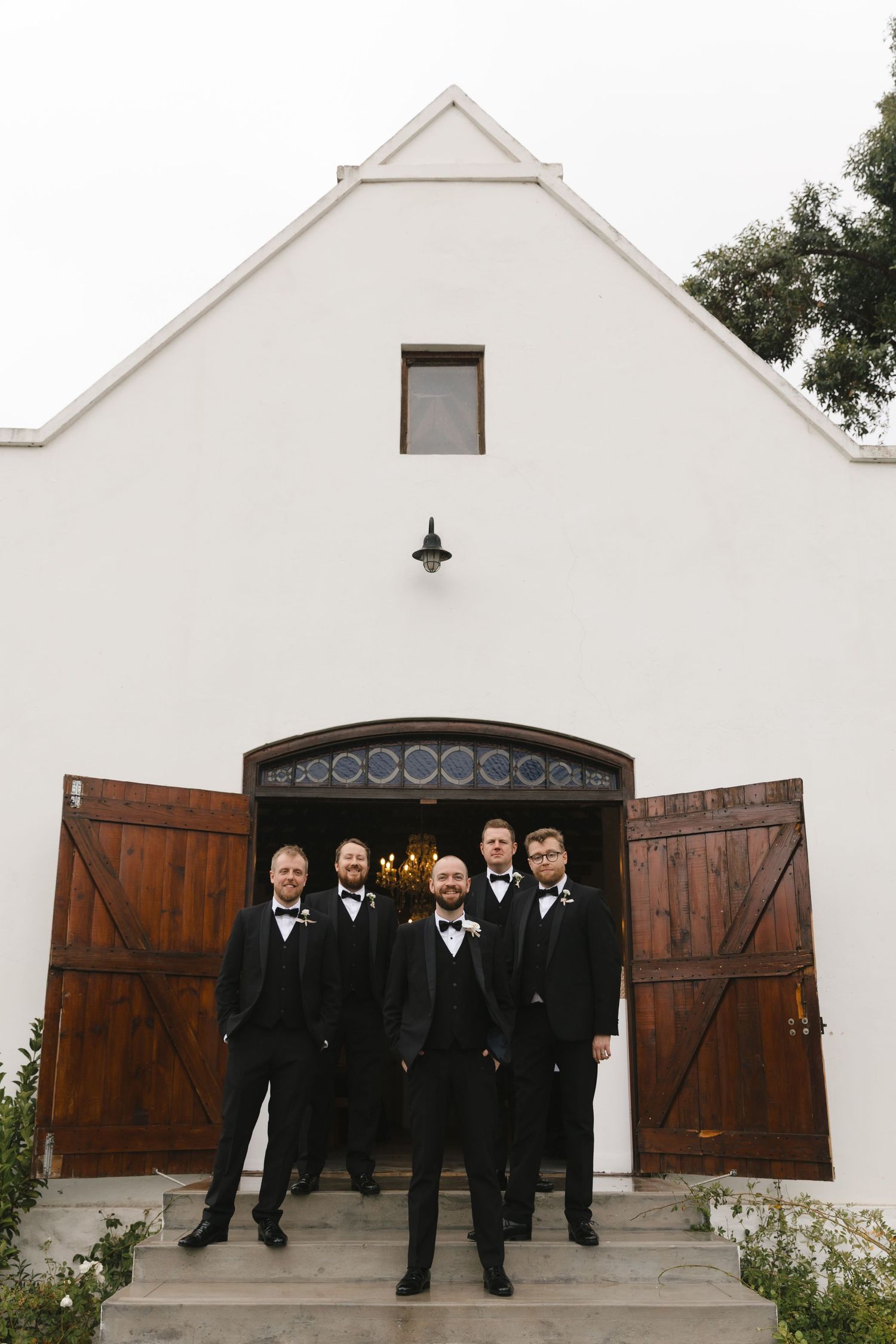 Wedding party members in formal black attire stand in front of white stucco building with wooden doors.