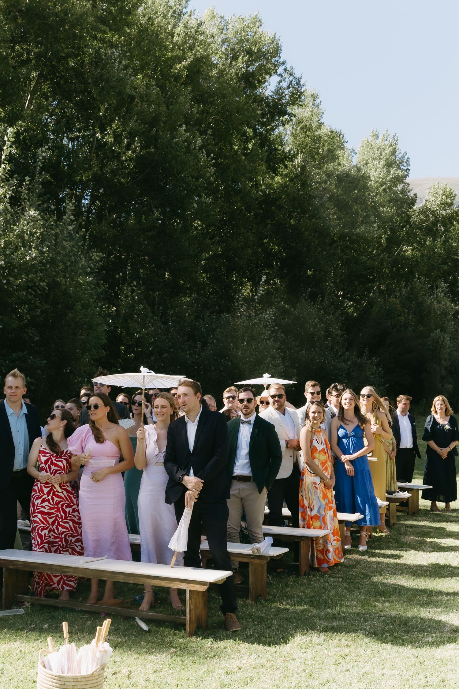 Guests gather outdoors under shaded trees at a summer wedding ceremony with white benches and casual attire.