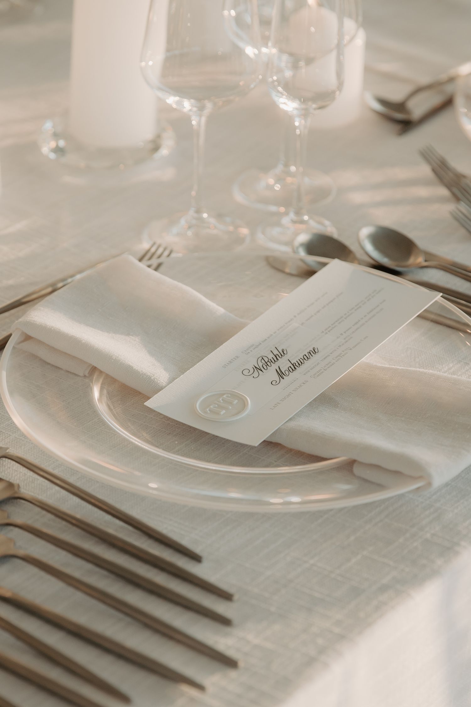 Elegant wedding place setting with white napkin, menu card, crystal glasses, and silver cutlery on linen tablecloth.