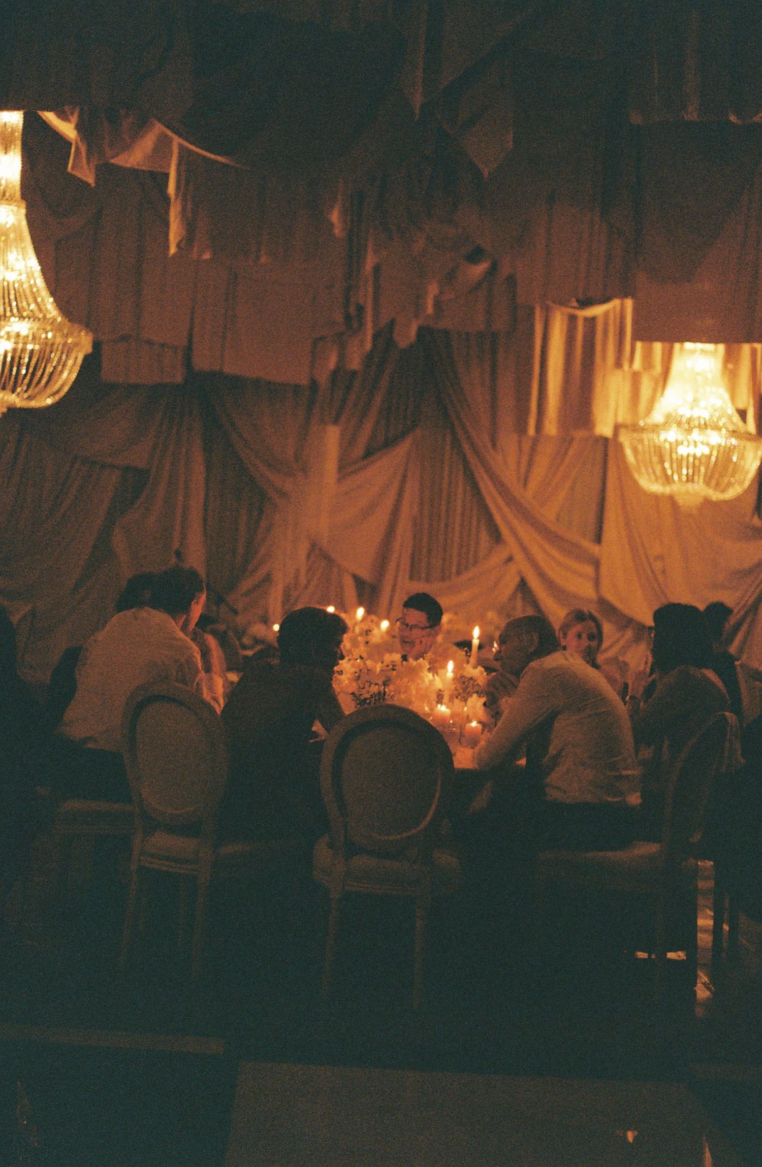 A dimly lit wedding reception venue with chandeliers and guests seated at long tables creating an intimate atmosphere.