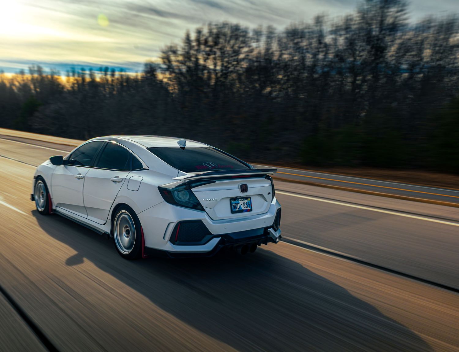 White Honda Civic Type R speeds down an empty highway during sunset with trees silhouetted in the background.