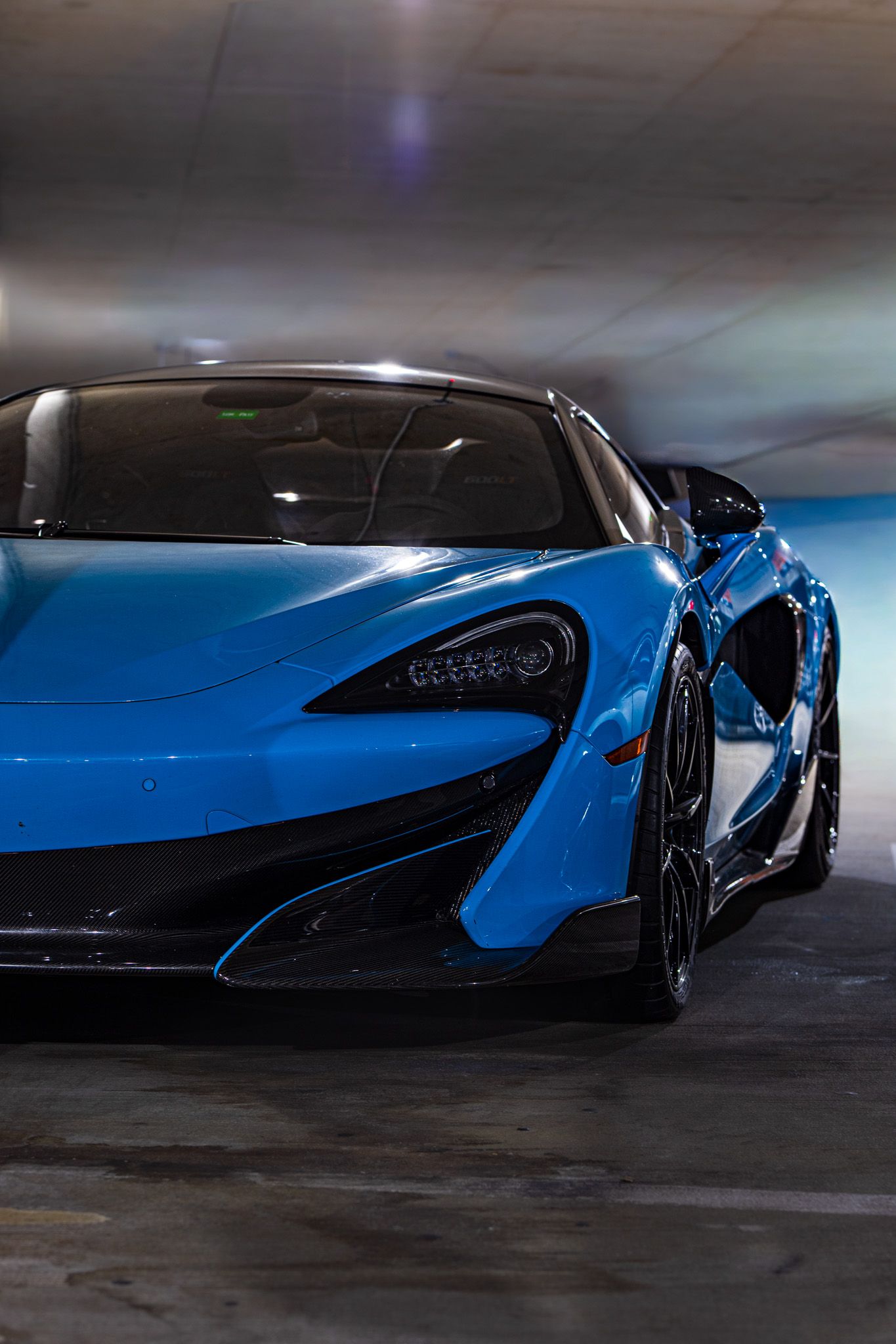 A sleek blue McLaren 600LT supercar with black accents parked in a dimly lit garage space.