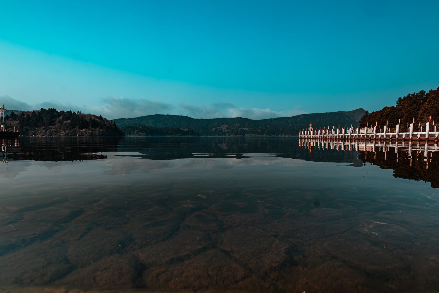 Serene mountain lake with mirror-like reflections and a dock at dusk under a clear blue sky.
