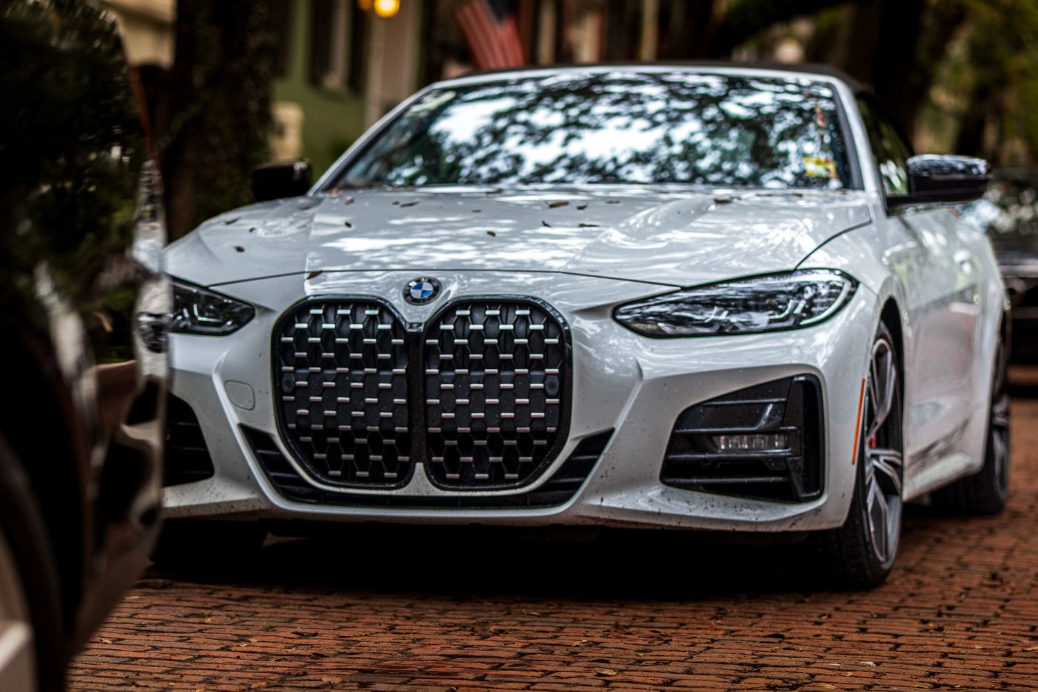 A white BMW M4 with distinctive large kidney grilles parked on a tree-lined street at dusk.