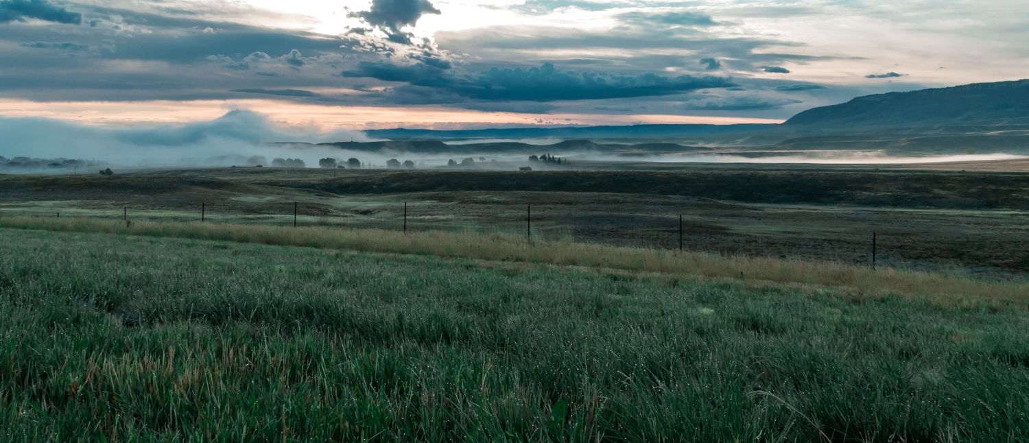 Panoramic view of a green meadow with rolling hills and dramatic cloudy skies during sunset.