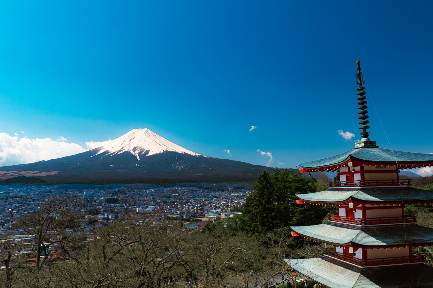 Mount Fuji with snow-capped peak behind red Chureito Pagoda and city of Fujiyoshida under clear blue sky.