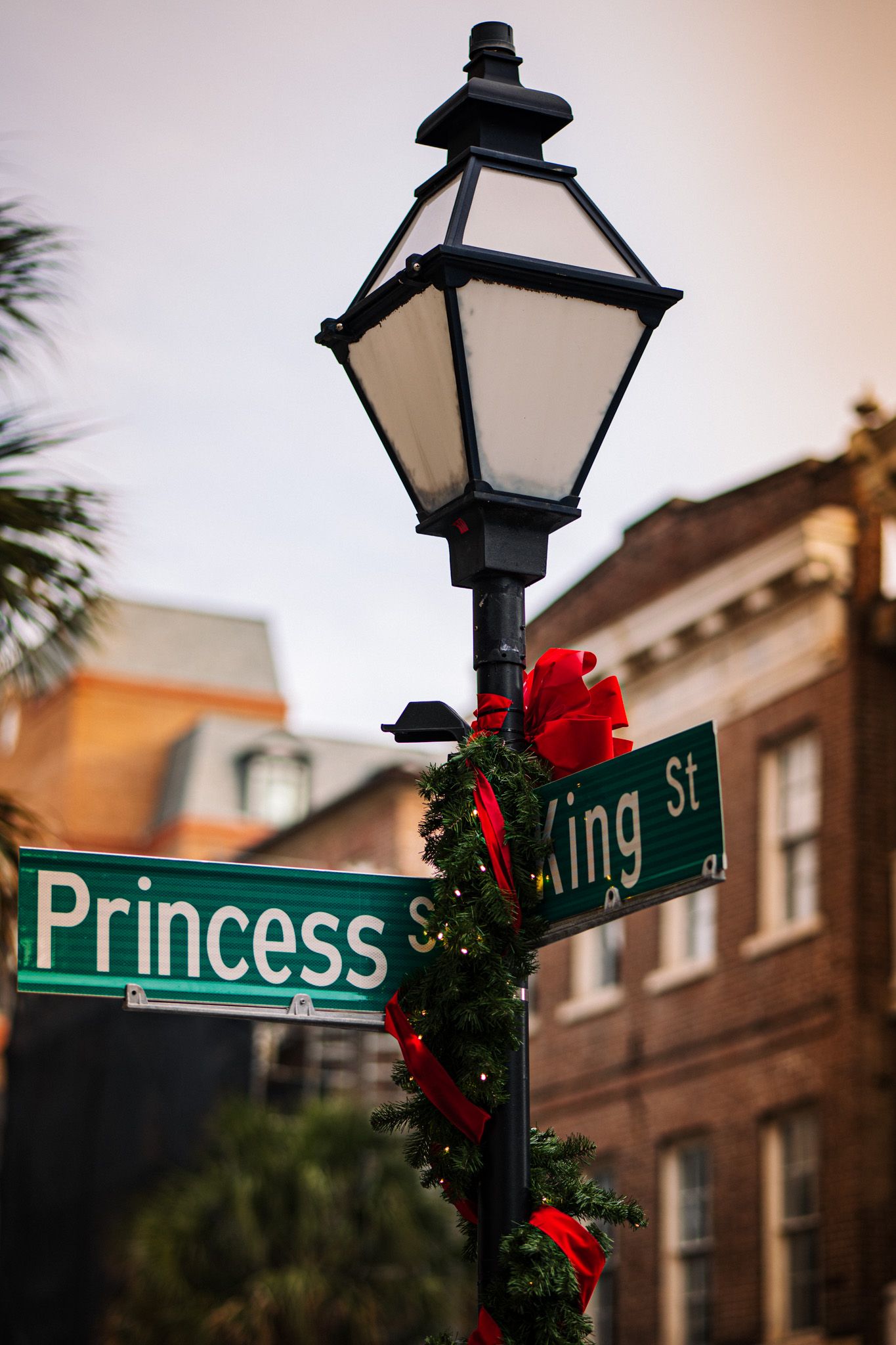 Street signs decorated with holiday wreaths and red bows on lamp posts in a historic downtown area at dusk.