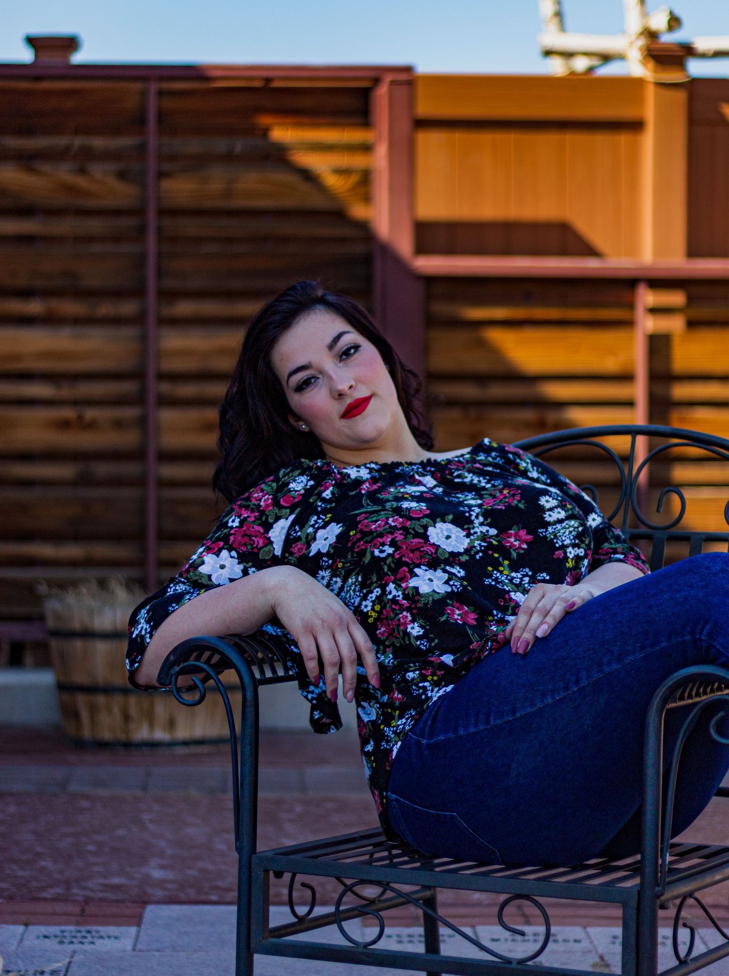 Woman in floral dress reclines on wrought iron bench during outdoor photoshoot.