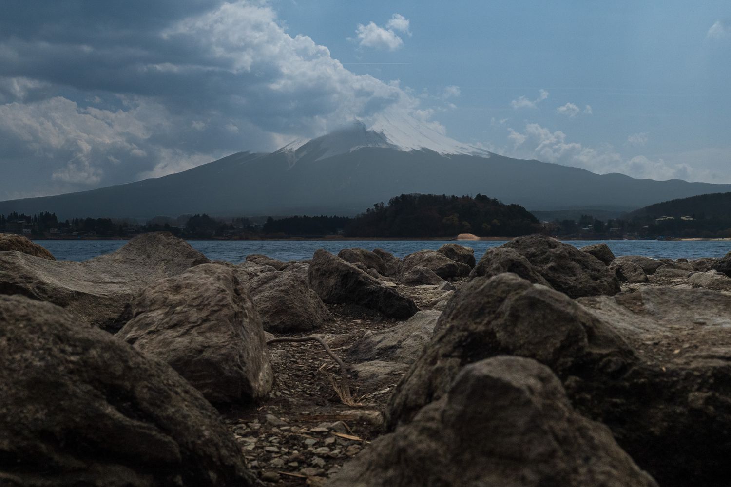 Rocky shoreline with dramatic mountain and cloudy sky reflected over a calm blue lake in Japan.