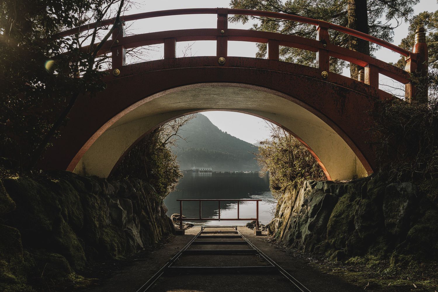 A red arched Japanese bridge framing a serene lake and mountain view through a stone tunnel passage.
