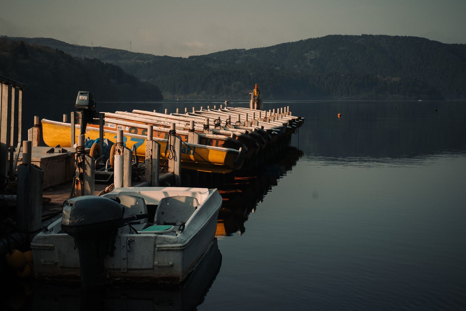 Boats docked at a calm lake pier during golden hour with misty mountains in the background.