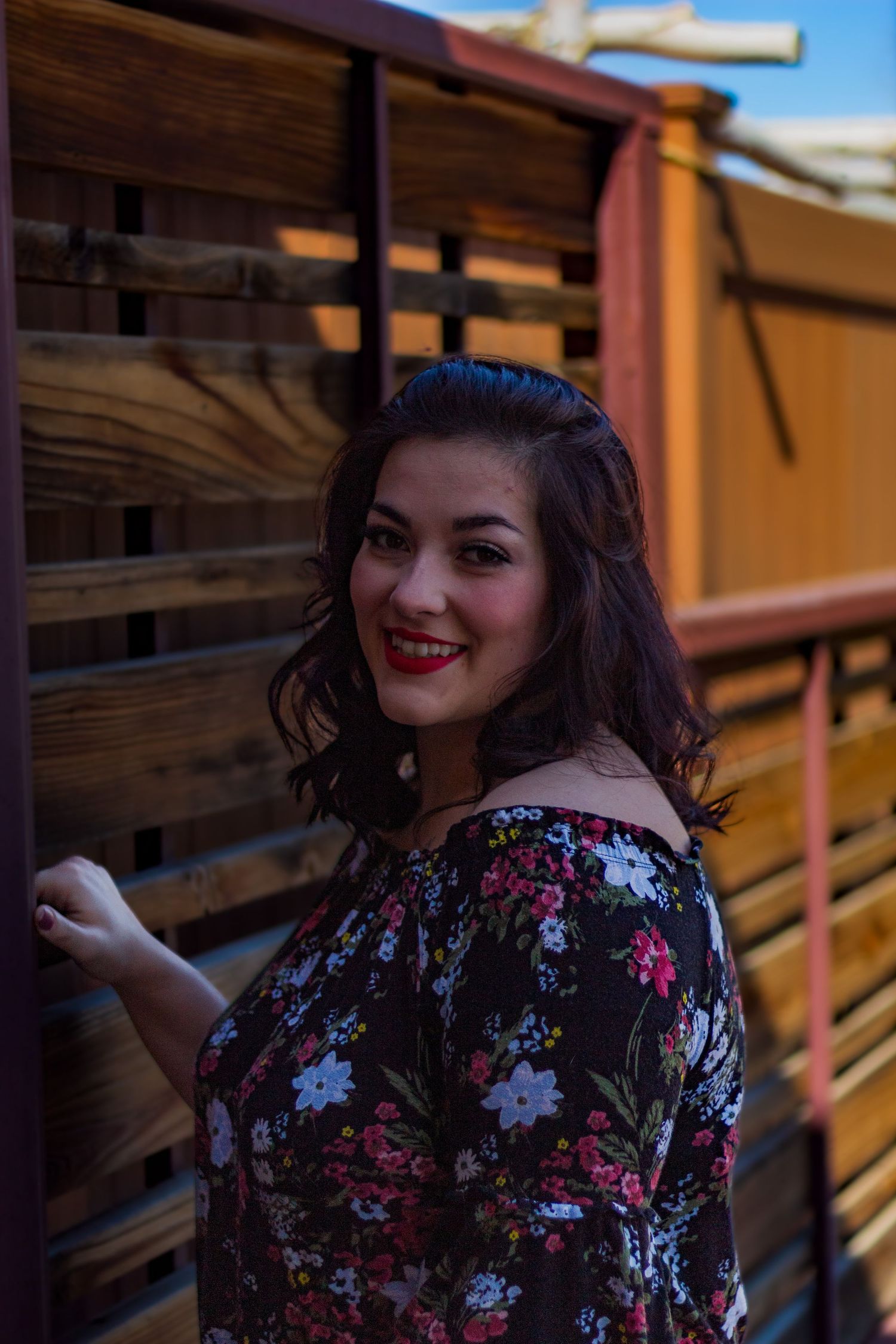 Woman in floral print dress poses against wooden backdrop with warm lighting and vintage styling.