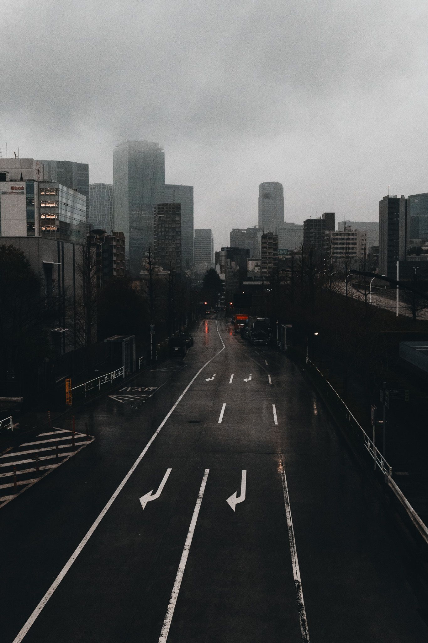 Moody rainy day on an empty urban road with white arrow markings leading to a foggy city skyline.