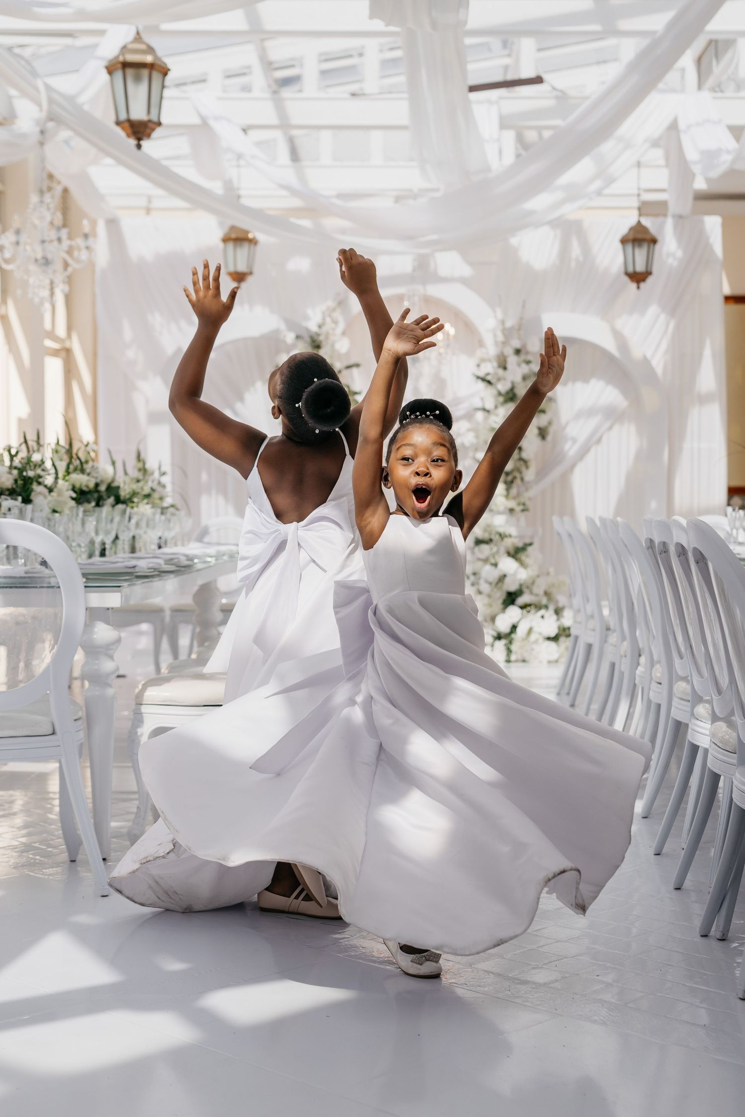 Two people in flowing white dresses dance joyfully at an elegant wedding venue decorated with lanterns and white draping.