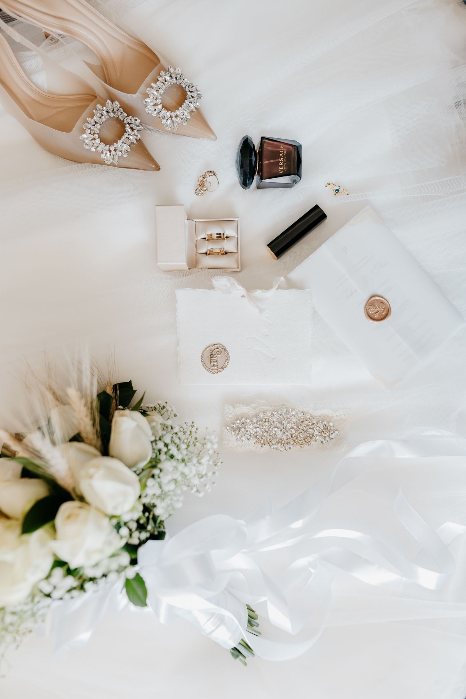 Wedding accessories including jewelry, perfume, and white floral arrangements elegantly arranged on white fabric.
