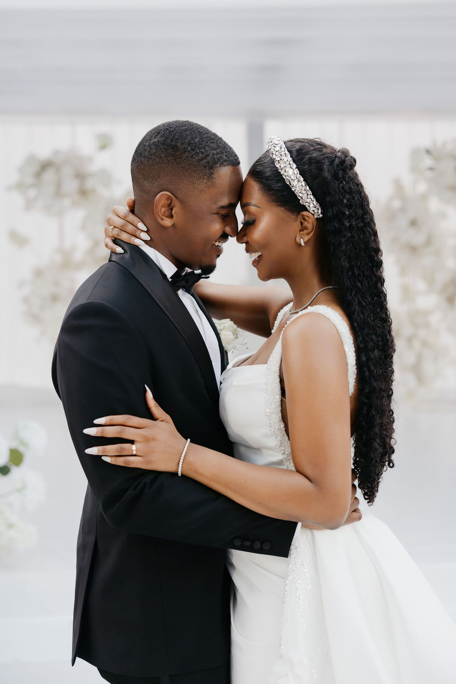 Romantic wedding couple embraces in an intimate moment against white floral backdrop.