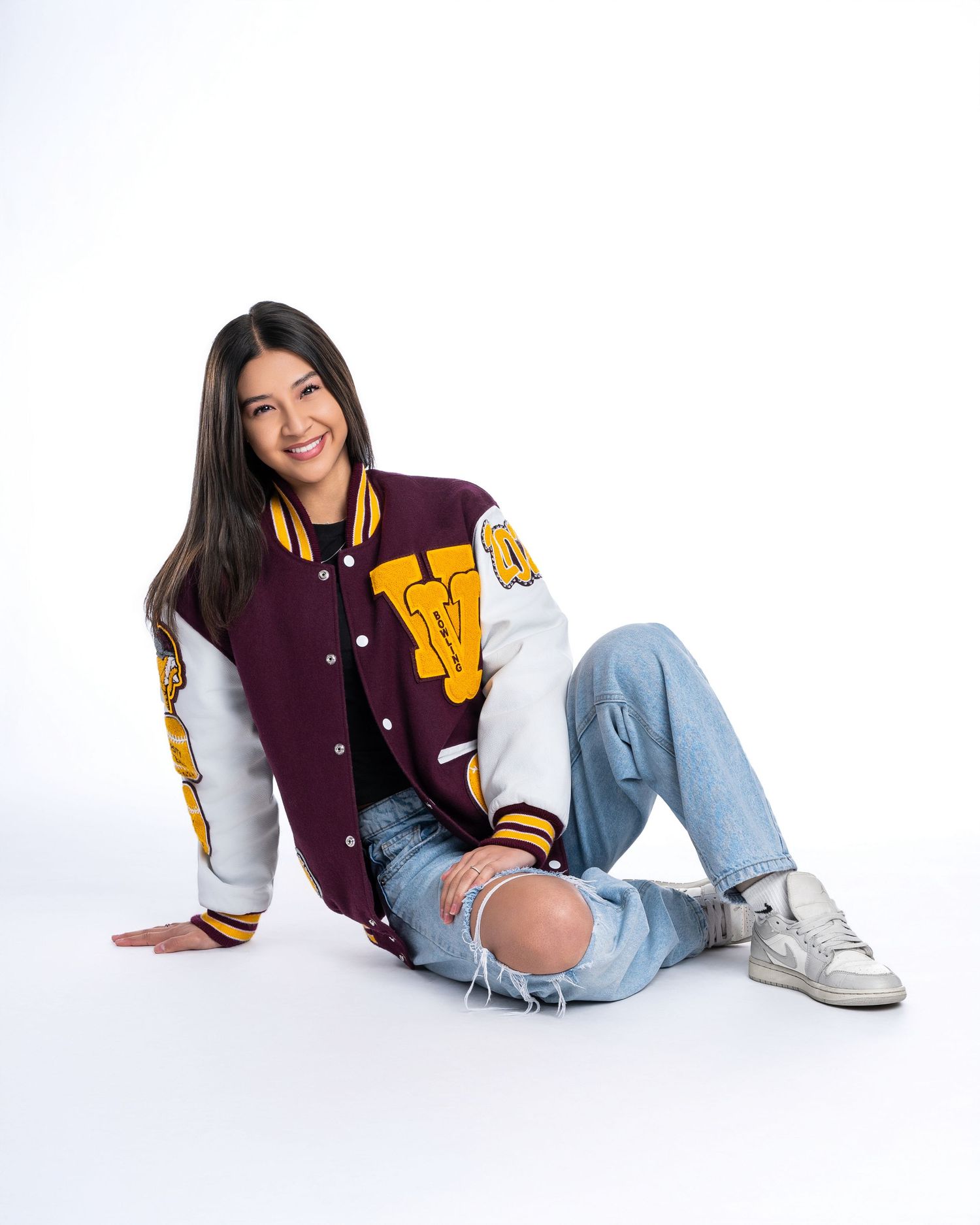 Smiling young woman in maroon and white varsity letterman jacket sitting on a white studio floor.