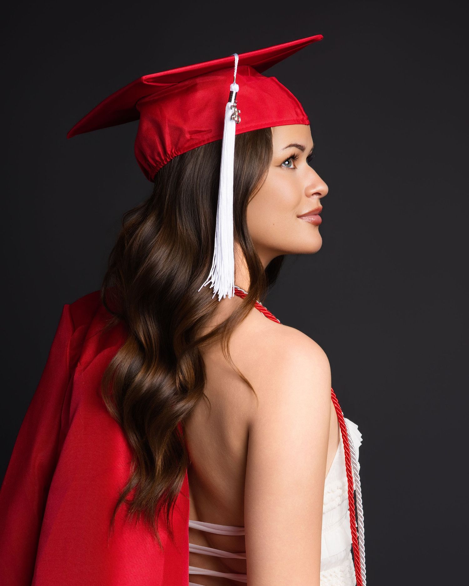 Female graduate in red cap and gown photographed from behind showing long brunette hair.