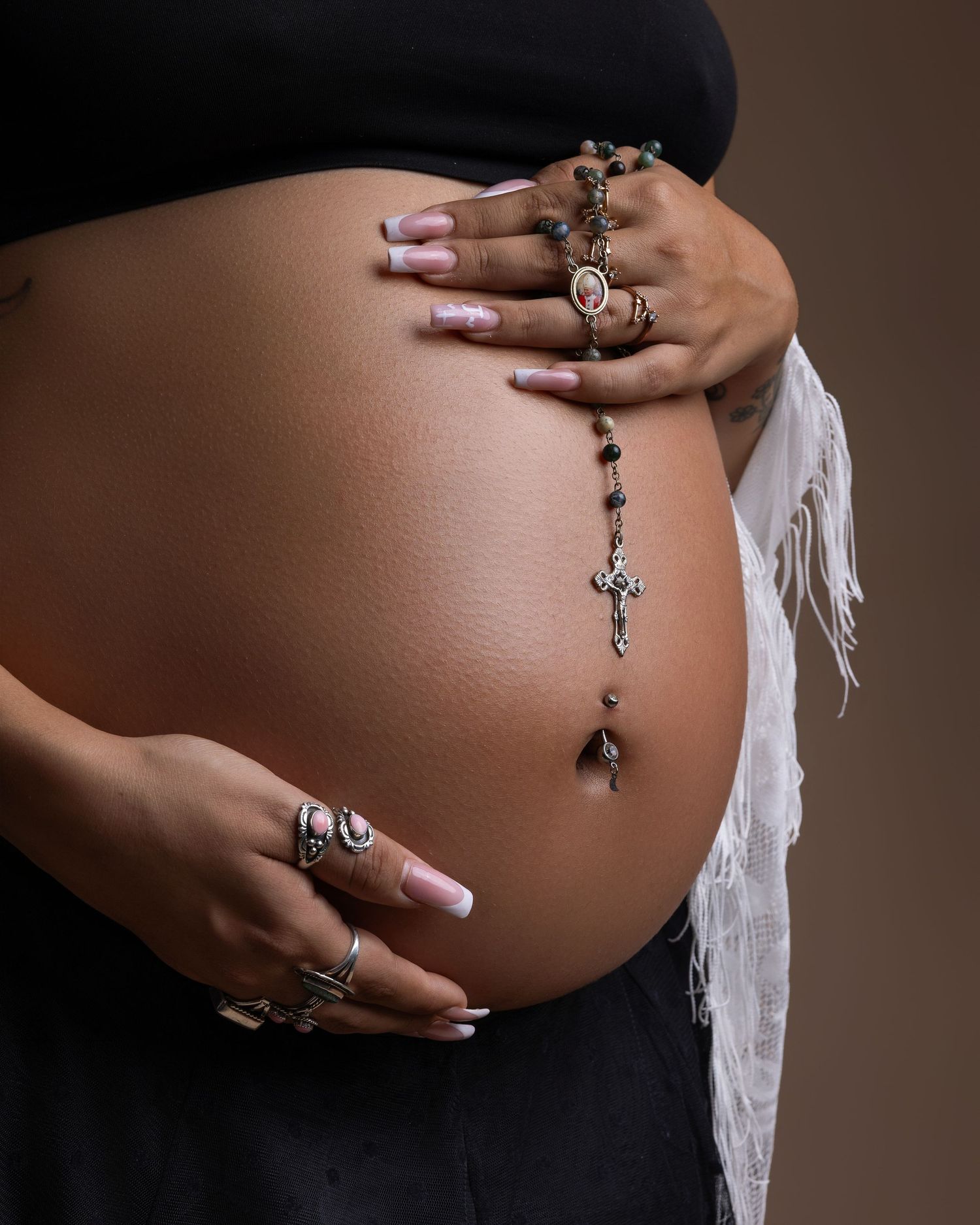 Close-up of pregnant belly with rosary beads, rings, and white fringe fabric on brown background.