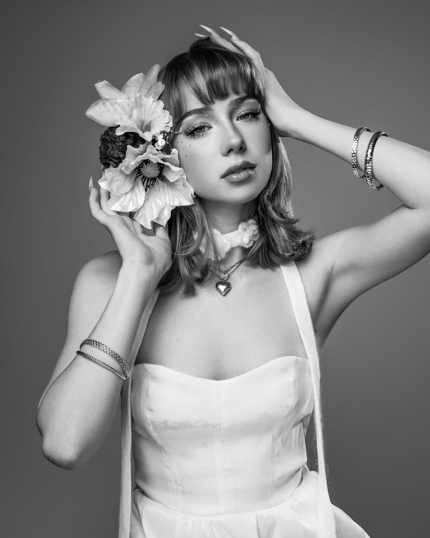 Black and white portrait of a young woman in white dress holding flowers, wearing heart necklace and bracelets.