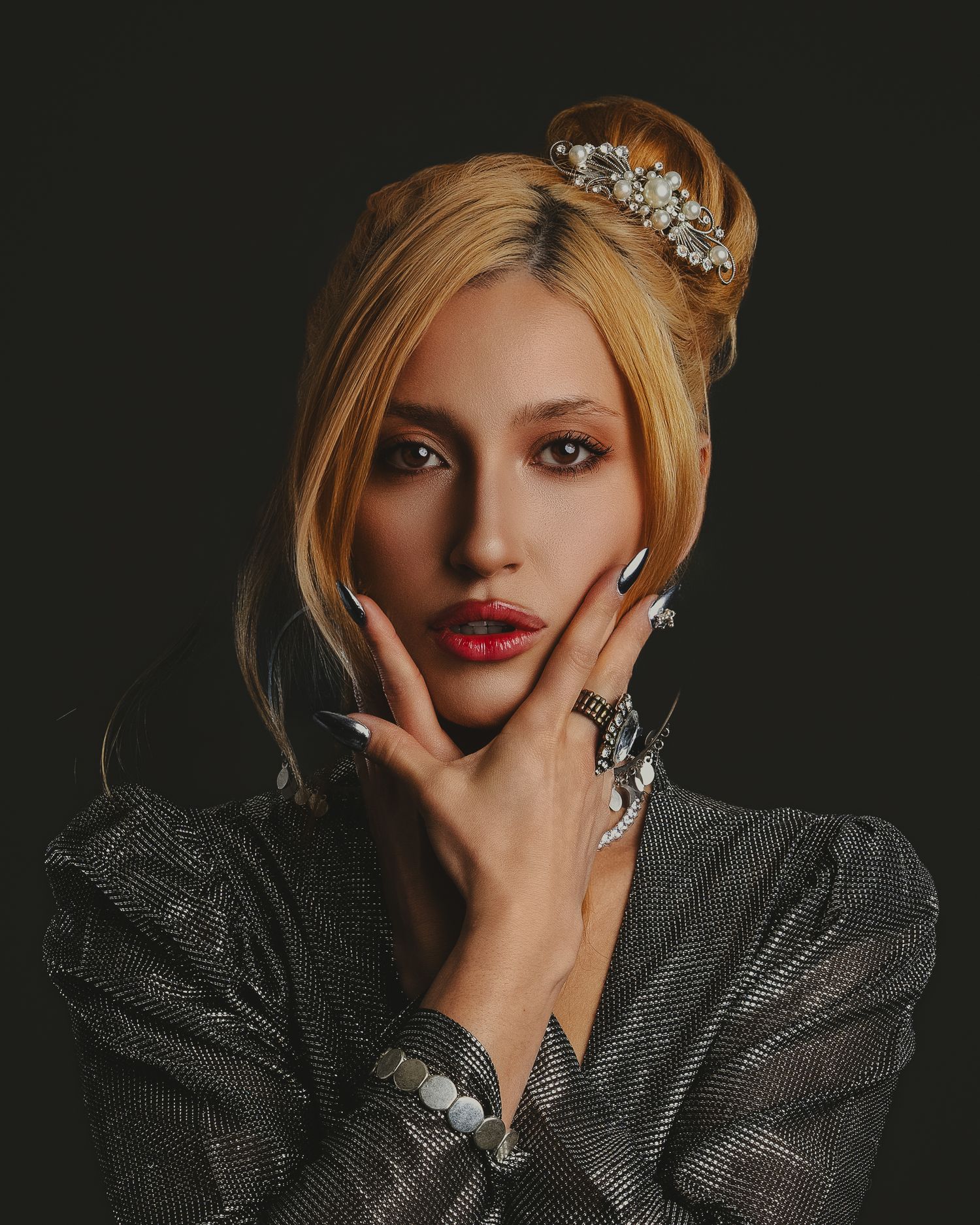 Blonde woman with crystal hair accessory, red lips, long nails, and rings poses elegantly in a silver blazer.
