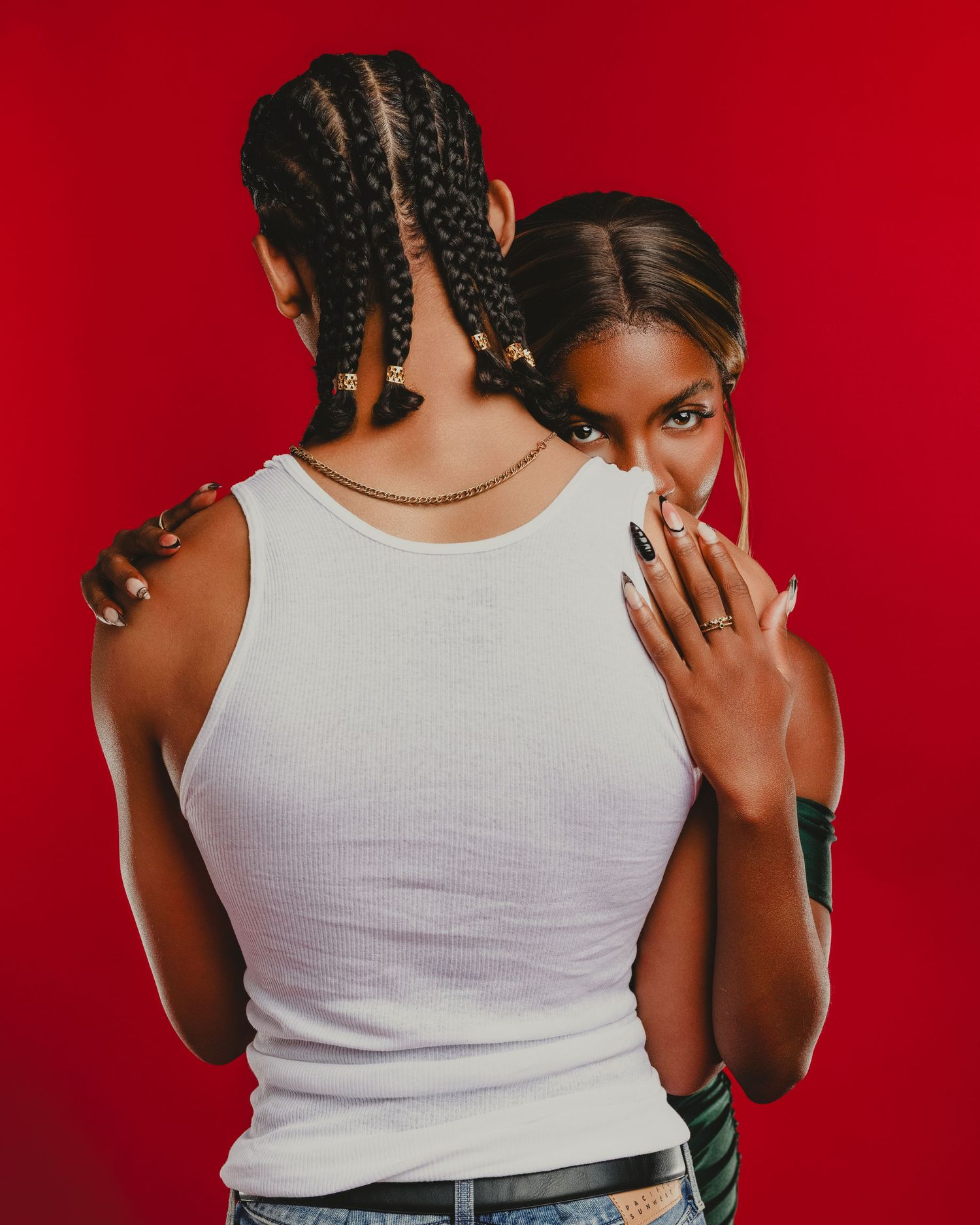Two Black women pose against a red background, one in a white tank top with braids, the other peering over her shoulder.