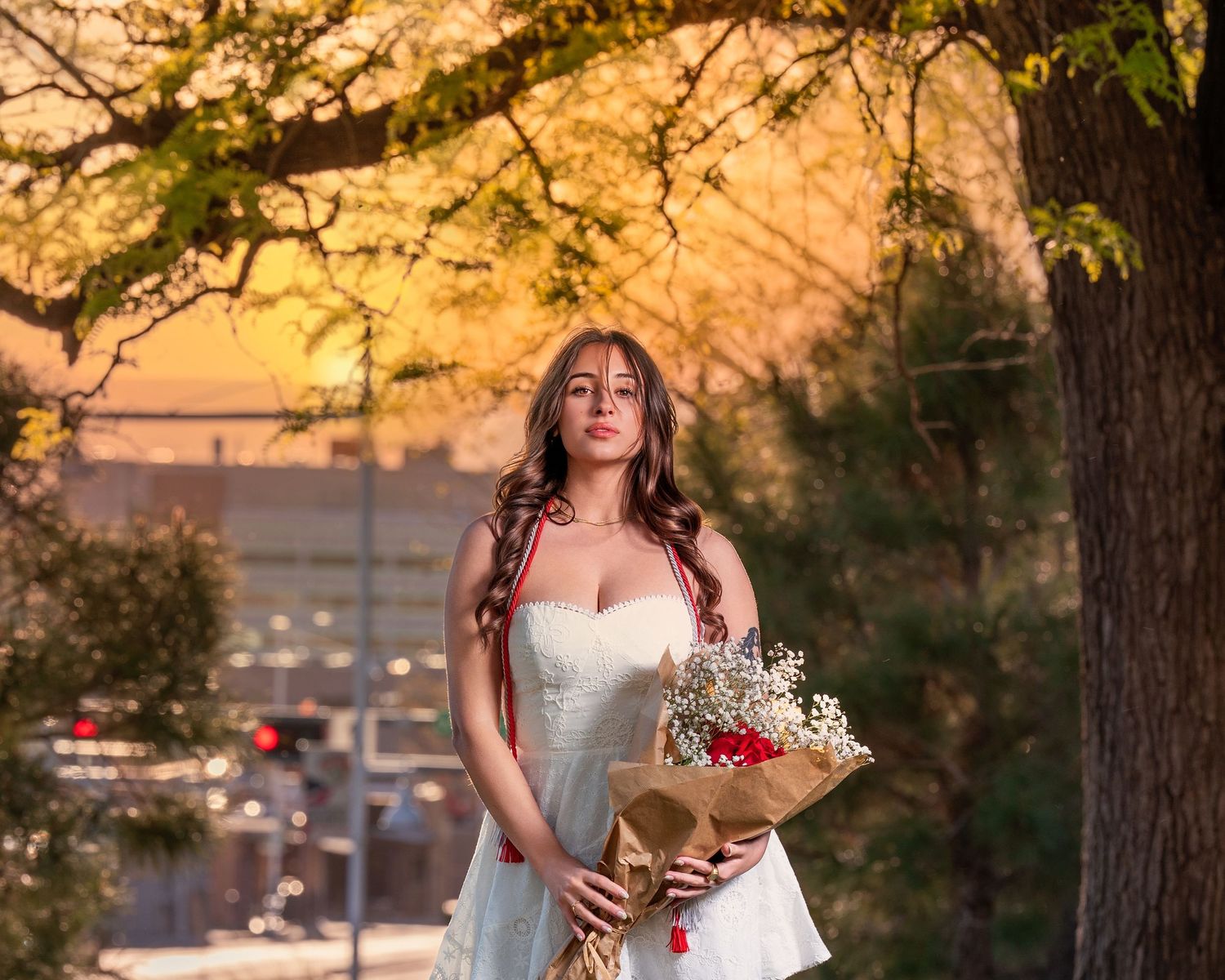 Young woman in white dress holds flower bouquet under golden sunset tree on city street.