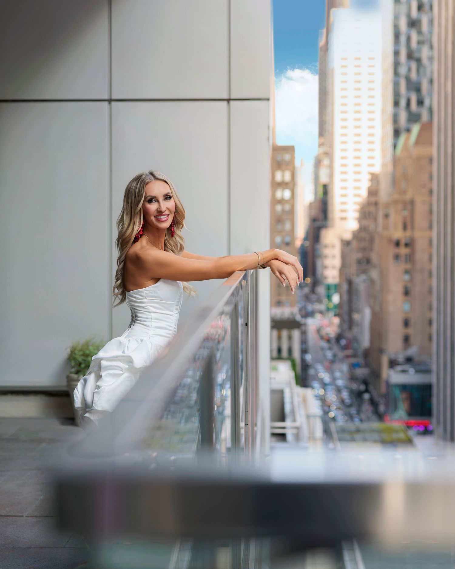 Smiling blonde woman in white dress posing on a city balcony with glass railing and NYC skyline backdrop.