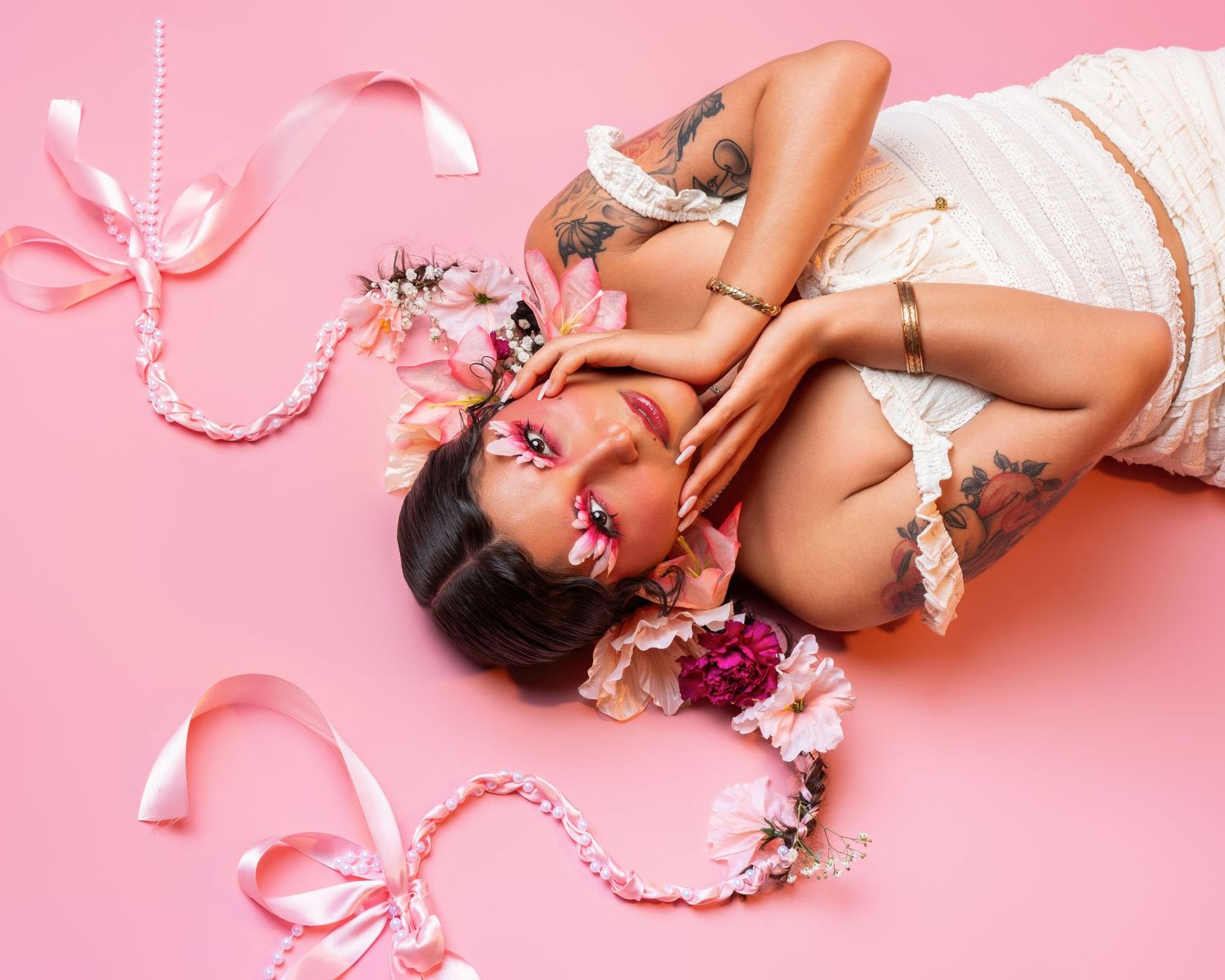 Tattooed woman lying on pink background surrounded by pink ribbons and floral decorations with artistic makeup.