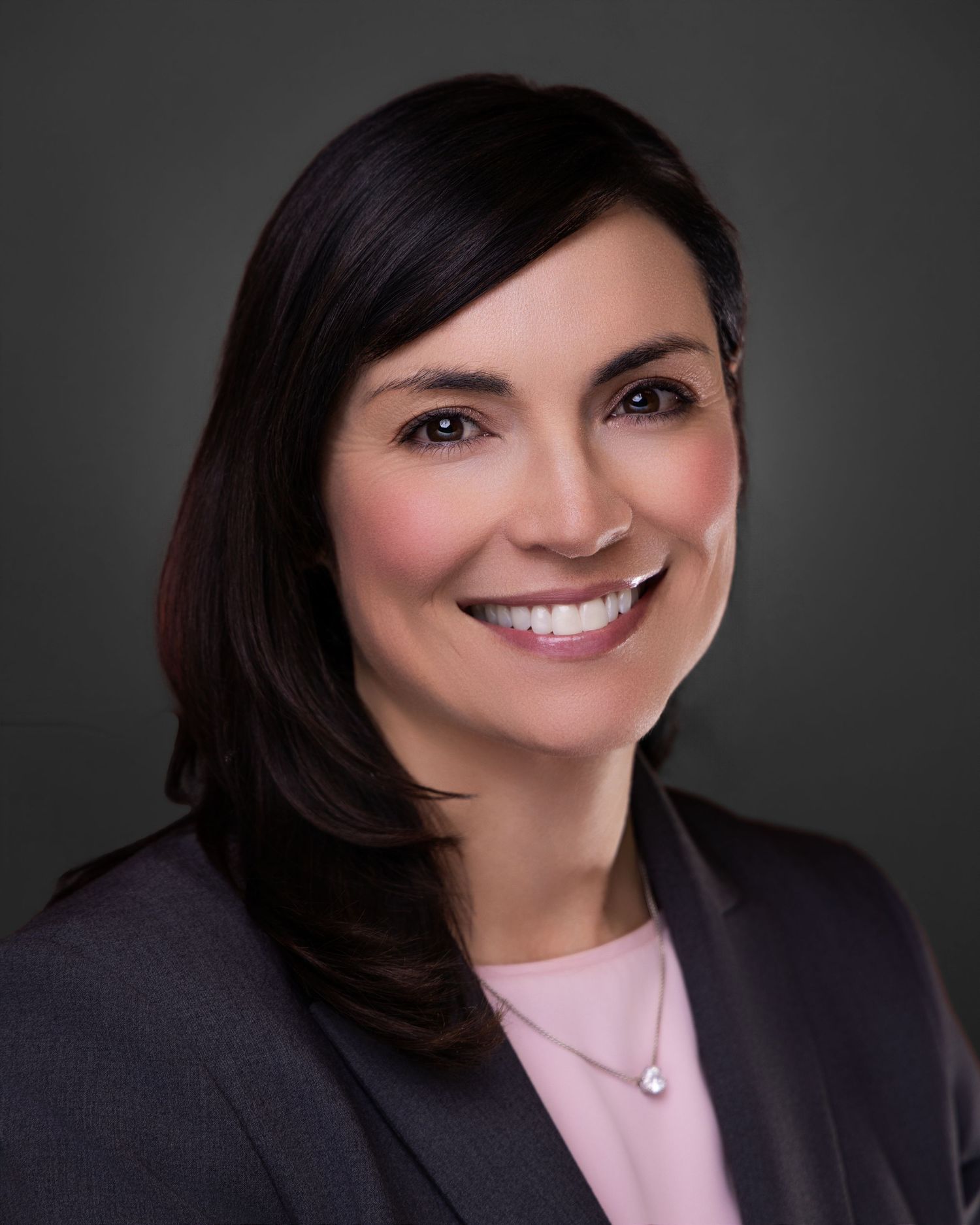 Professional headshot of smiling brunette businesswoman in gray blazer and pink top against dark background.