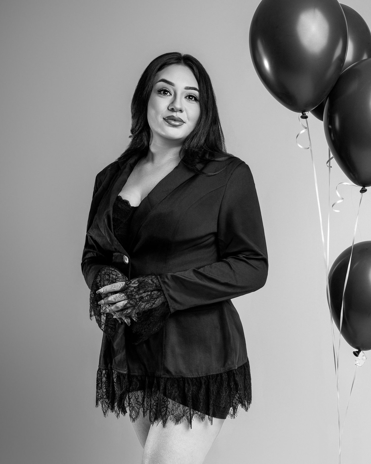 Black and white photo of a woman in a black blazer and lace skirt posing with dark balloons.