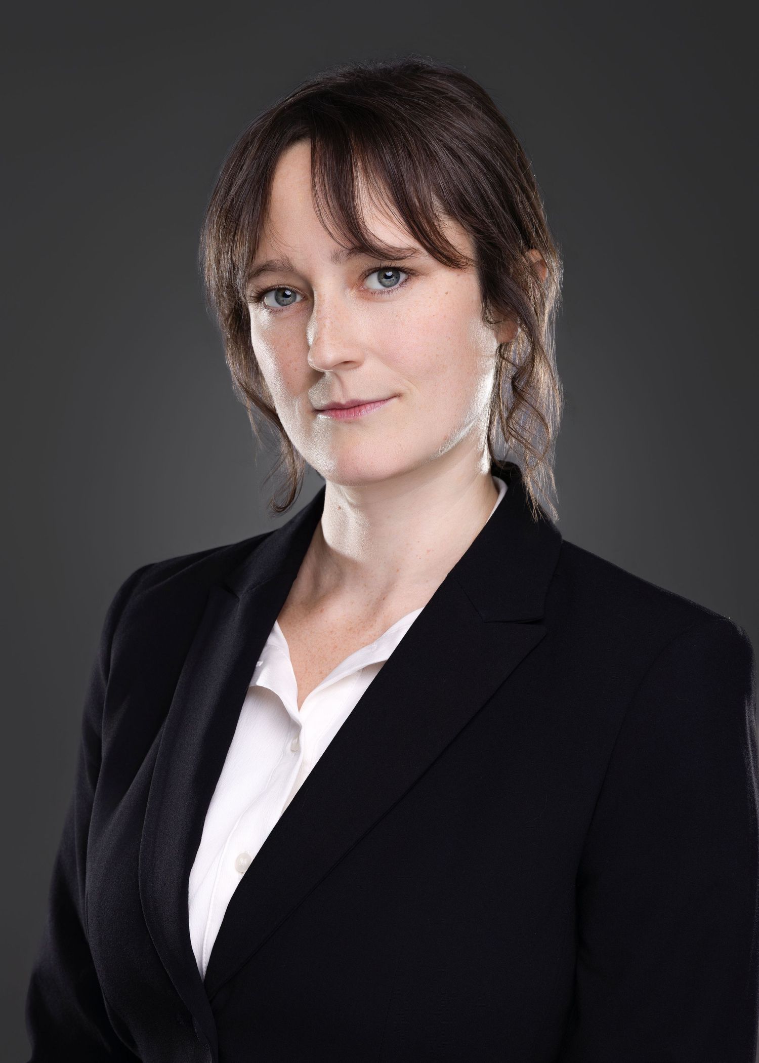 Professional headshot of serious young woman with updo hairstyle in black blazer and white shirt on gray background.