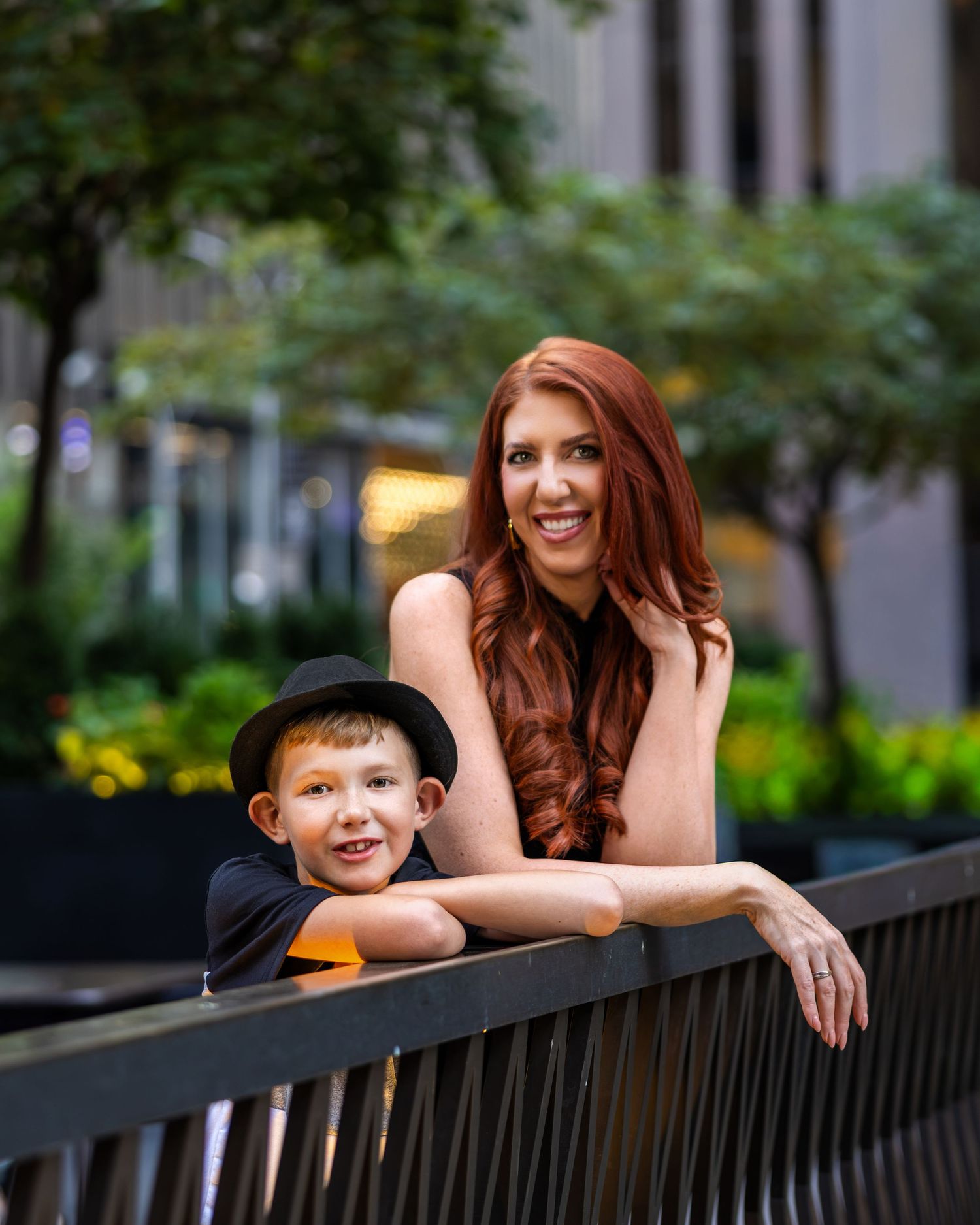 Smiling redhead woman and young boy in a black hat posing together on a bridge outdoors.