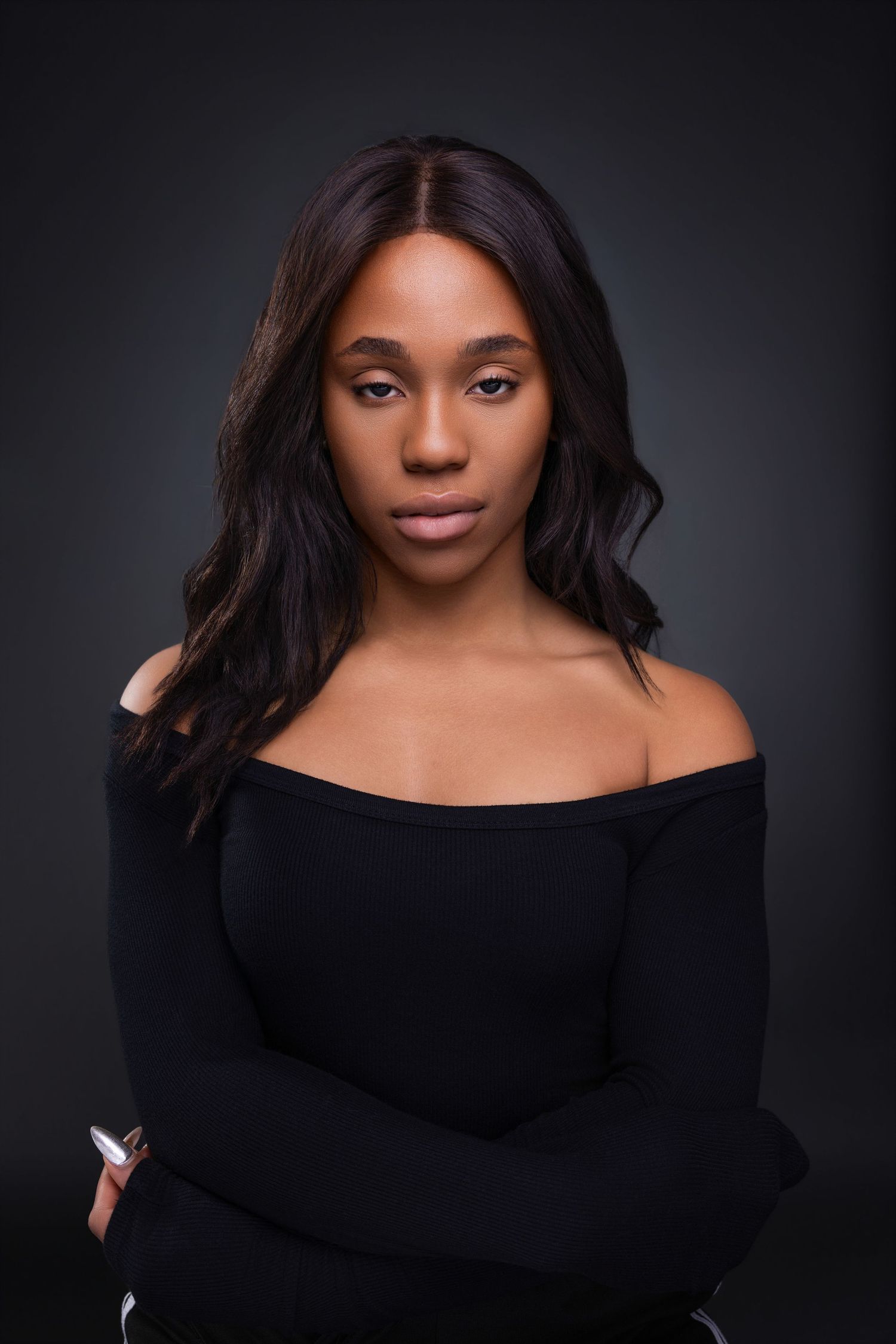 A confident young Black woman in an off-shoulder black top poses with arms crossed against a dark studio background.
