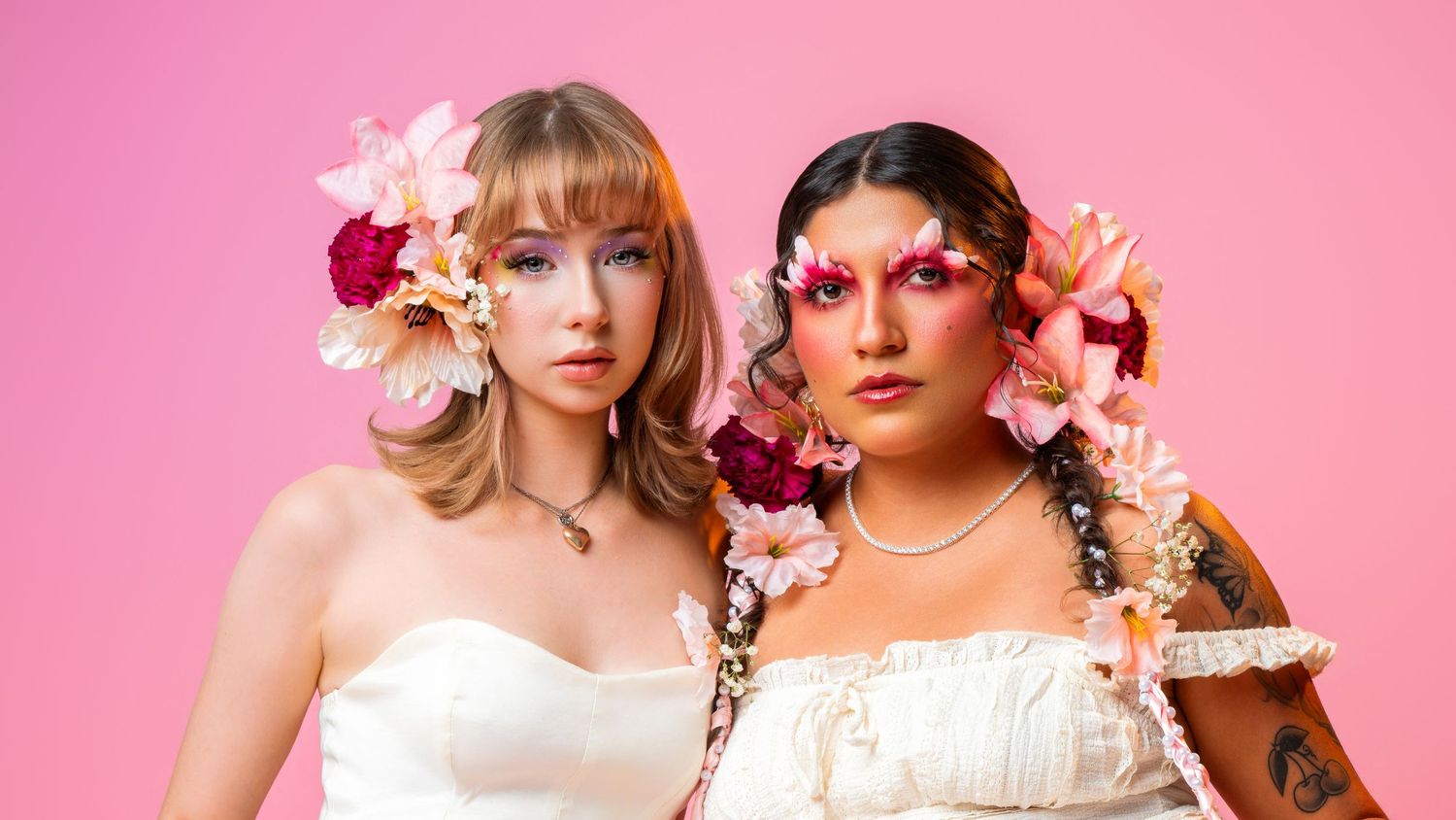 Two women in white dresses with floral hair accessories pose against a pink background.