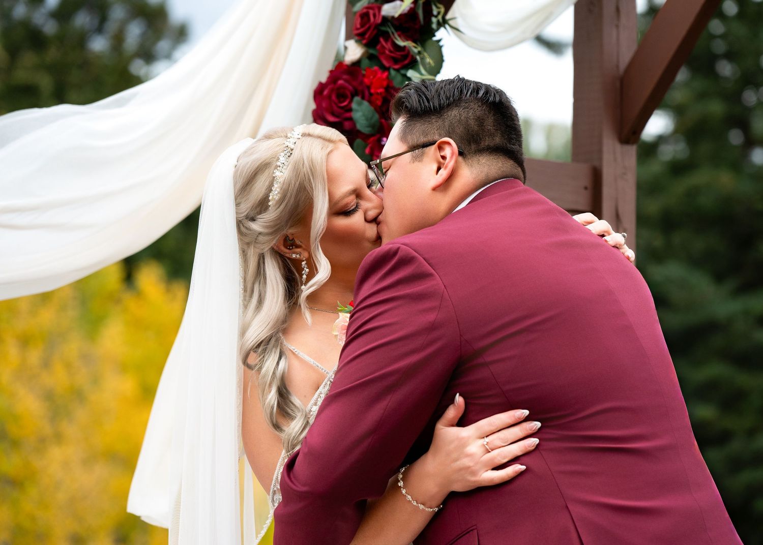 Bride and groom share their first kiss at outdoor fall wedding ceremony under floral arch with red roses.