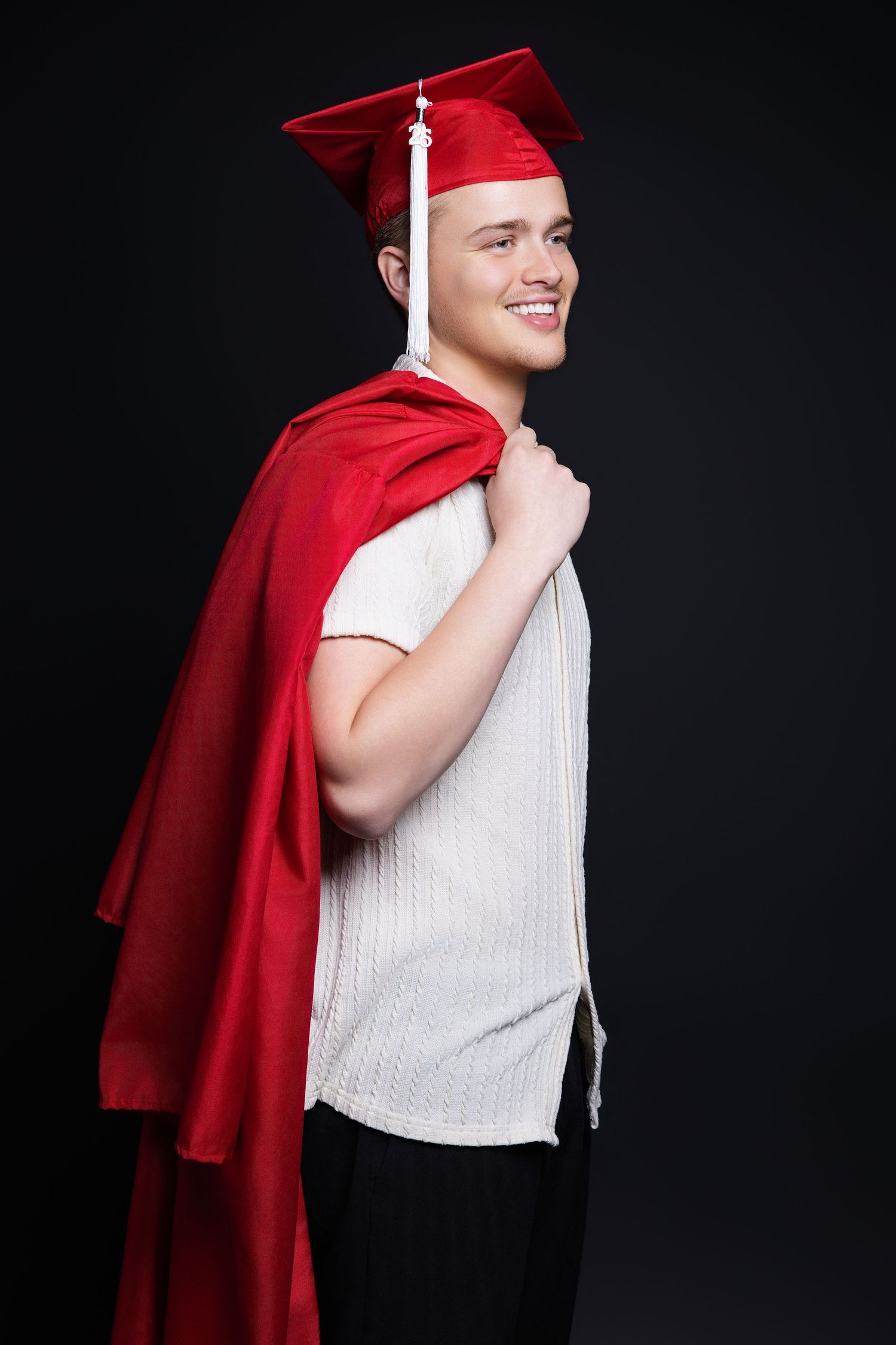 Young man in white shirt and red graduation cap and gown smiling in a dark studio setting.