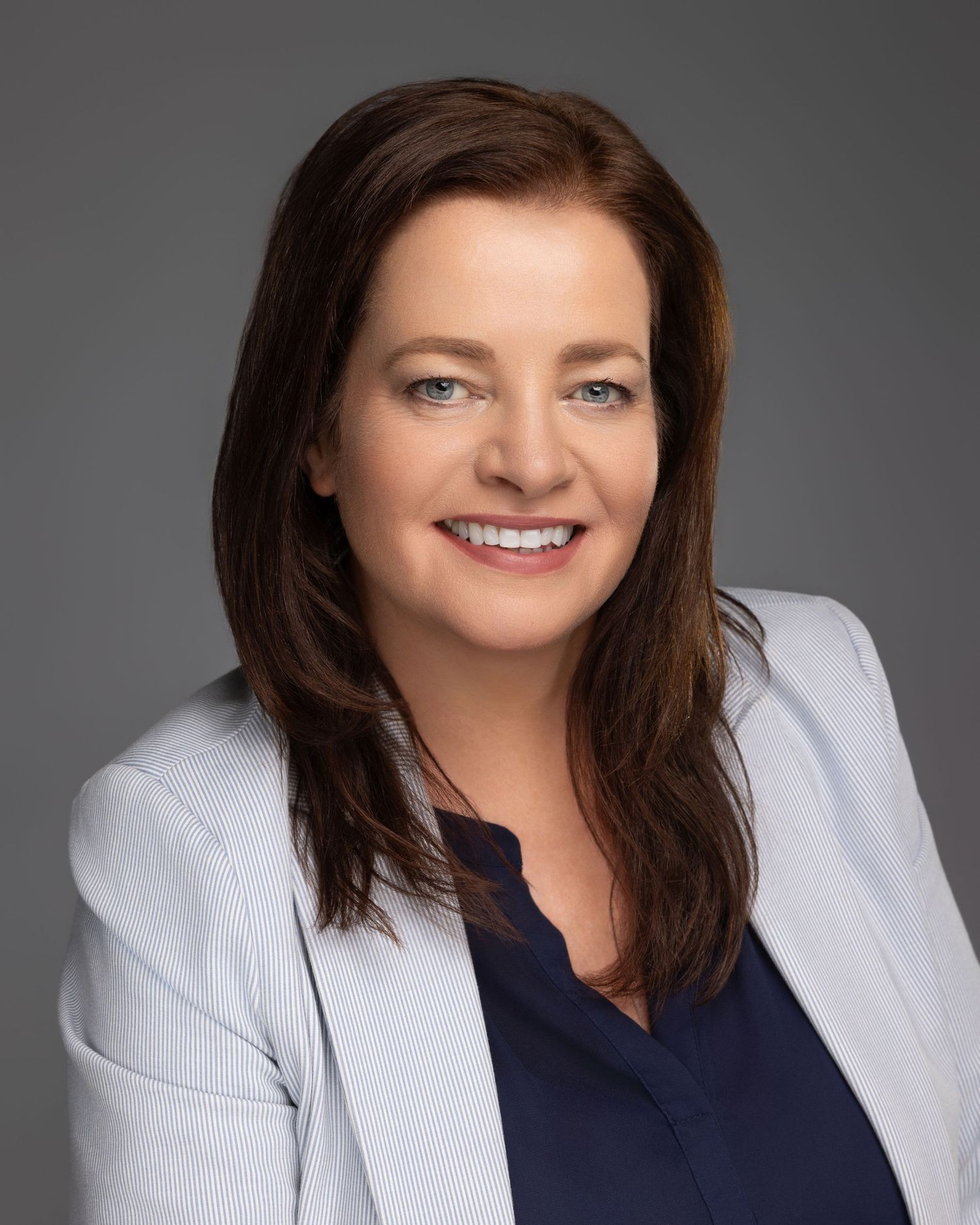 Professional headshot of smiling woman with brown hair in light gray blazer and navy top on gray background.