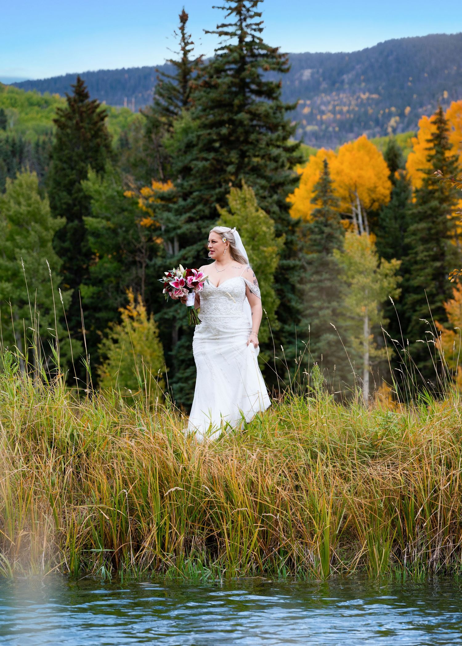 Bride in white gown holds pink bouquet standing by mountain lake with colorful fall foliage.