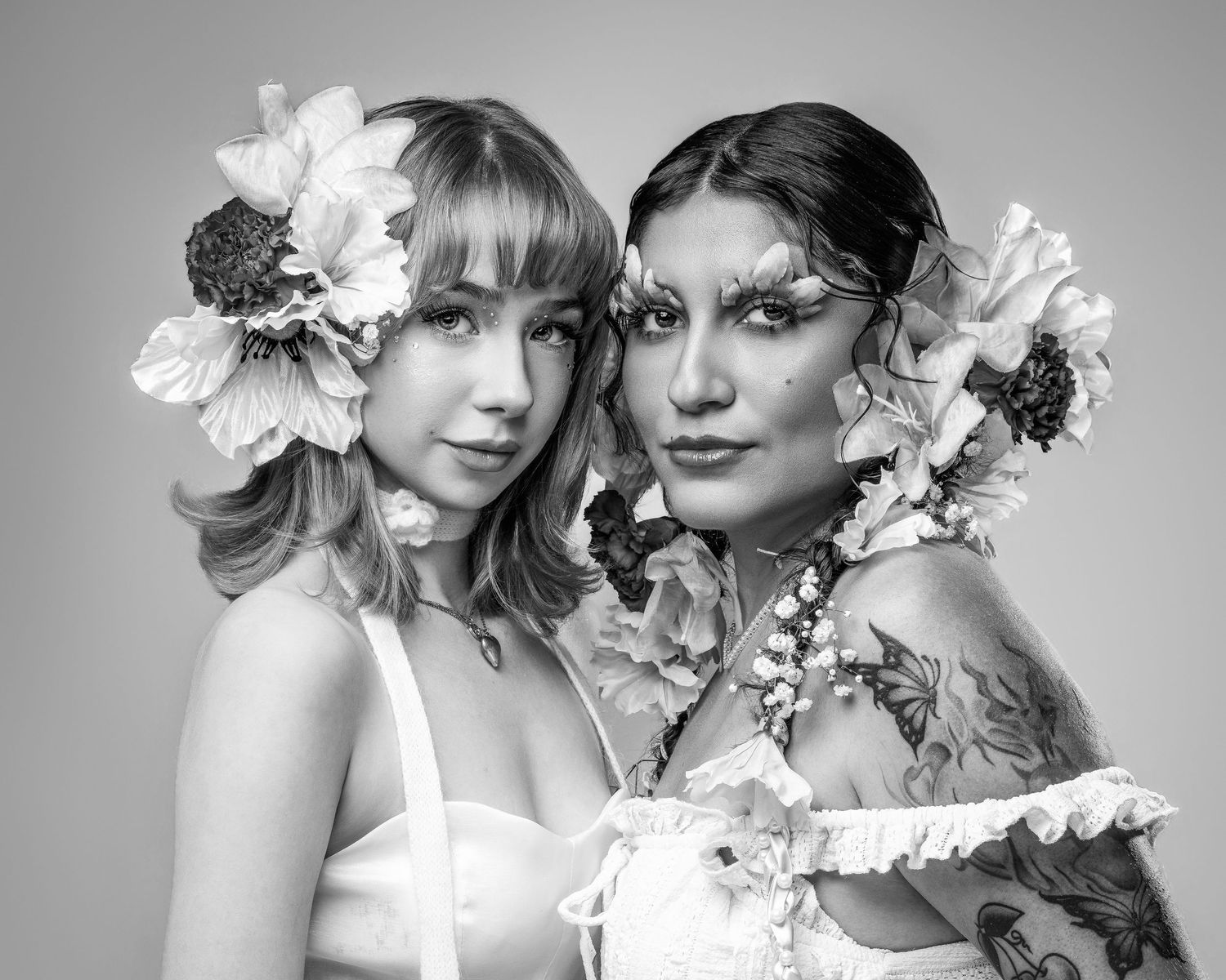 Two women with floral hair accessories pose together in a black and white portrait, one with tattoos and dramatic eye makeup.