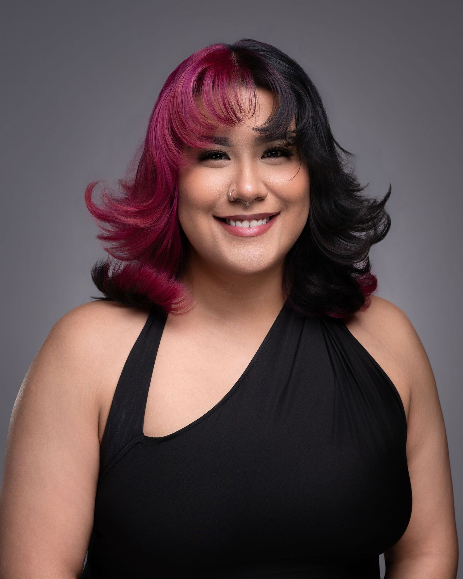 Woman with split pink and black hair smiling, wearing black one-shoulder top, studio portrait.