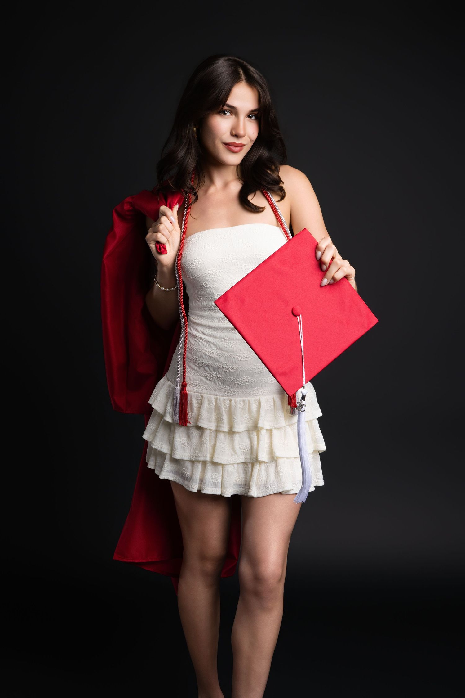 Young woman in white dress holding red graduation cap and gown in a dark studio setting.
