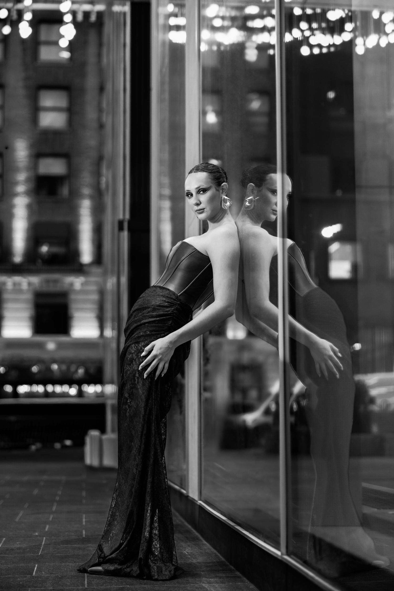 Black and white photo of an elegant woman in a backless gown leaning against a glass window on a city street at night.