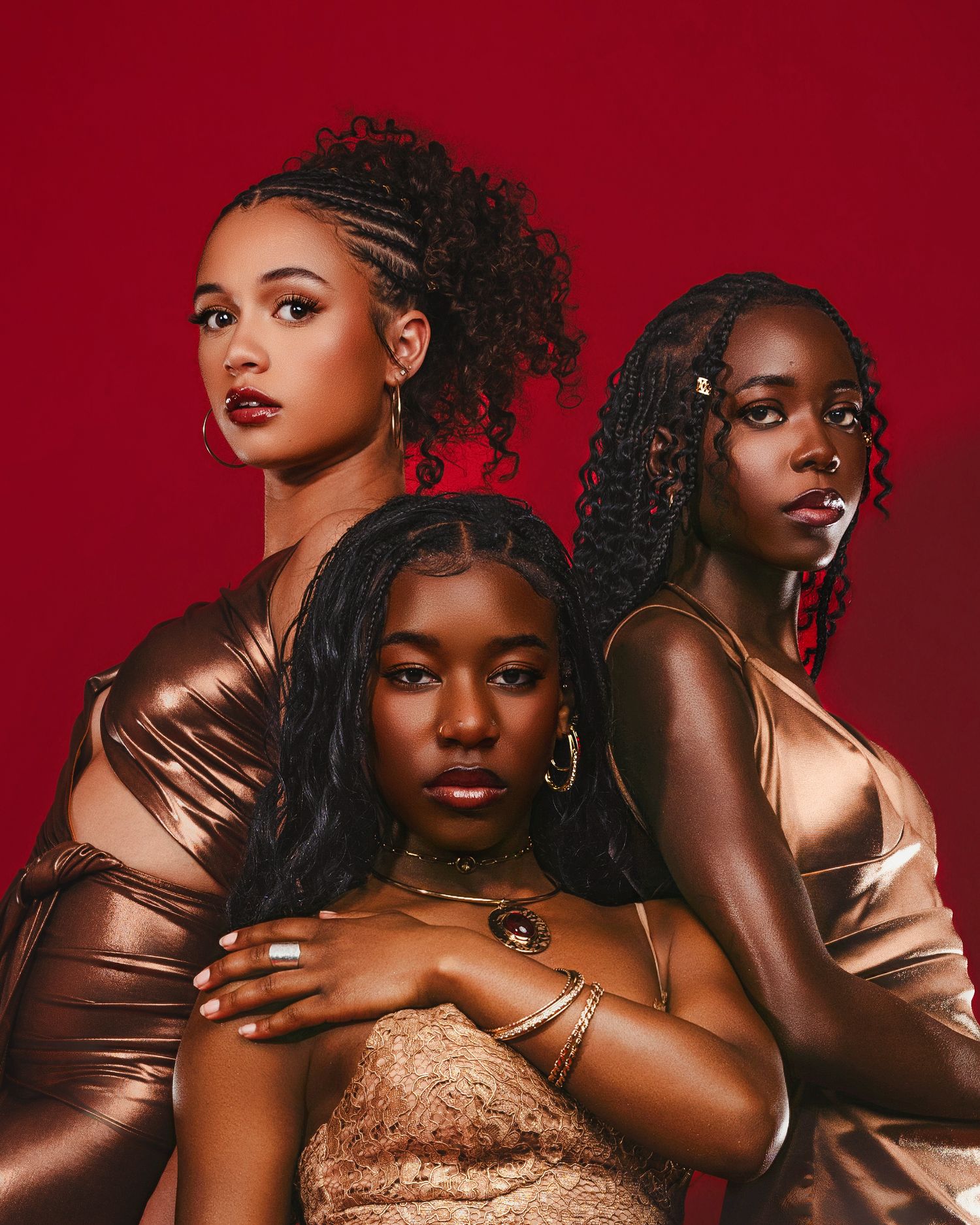 Three young women in bronze and gold outfits pose confidently against a bold red background.