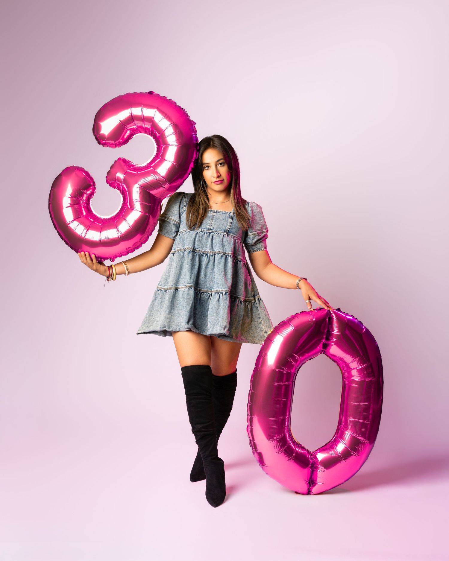Woman in blue dress and black boots holds pink number 30 balloons against a pink background.