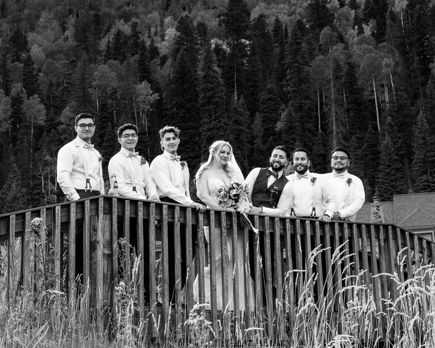 Wedding party posing on wooden bridge surrounded by lush forest in black and white photo.