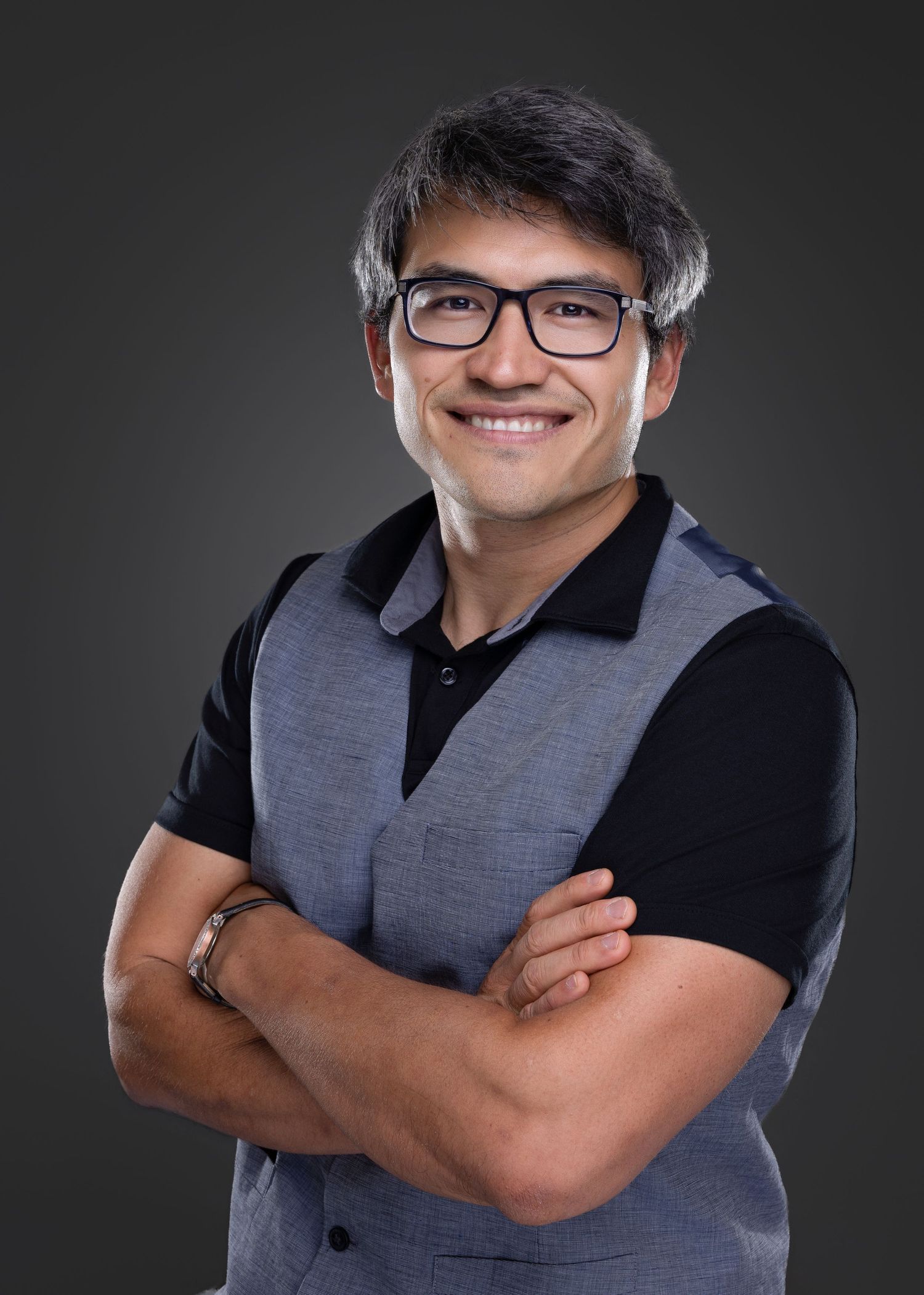 Friendly man with glasses wearing gray vest and black shirt smiling with arms crossed on dark background.