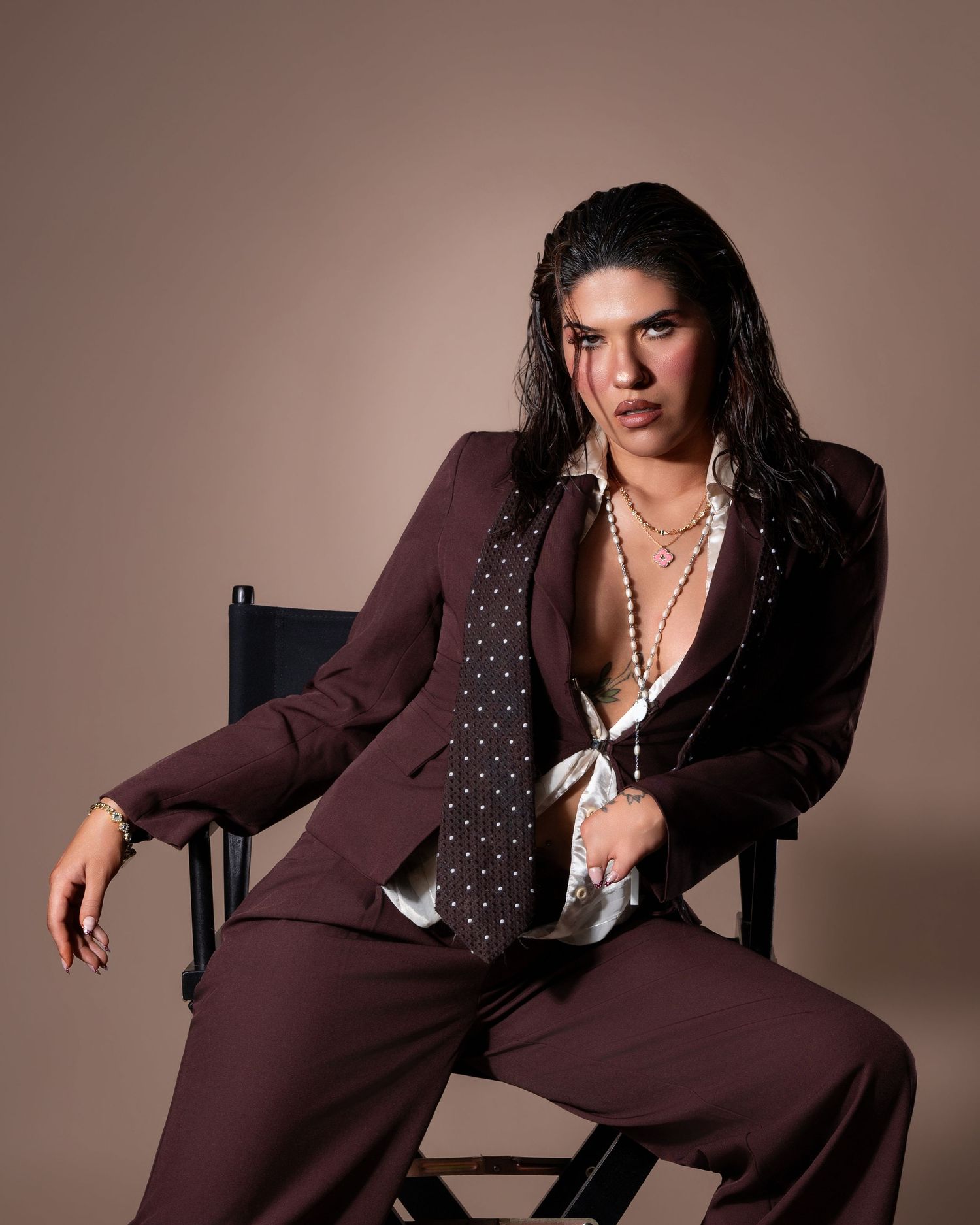 Person in brown polka dot suit with pearl necklace seated in director's chair against beige background.