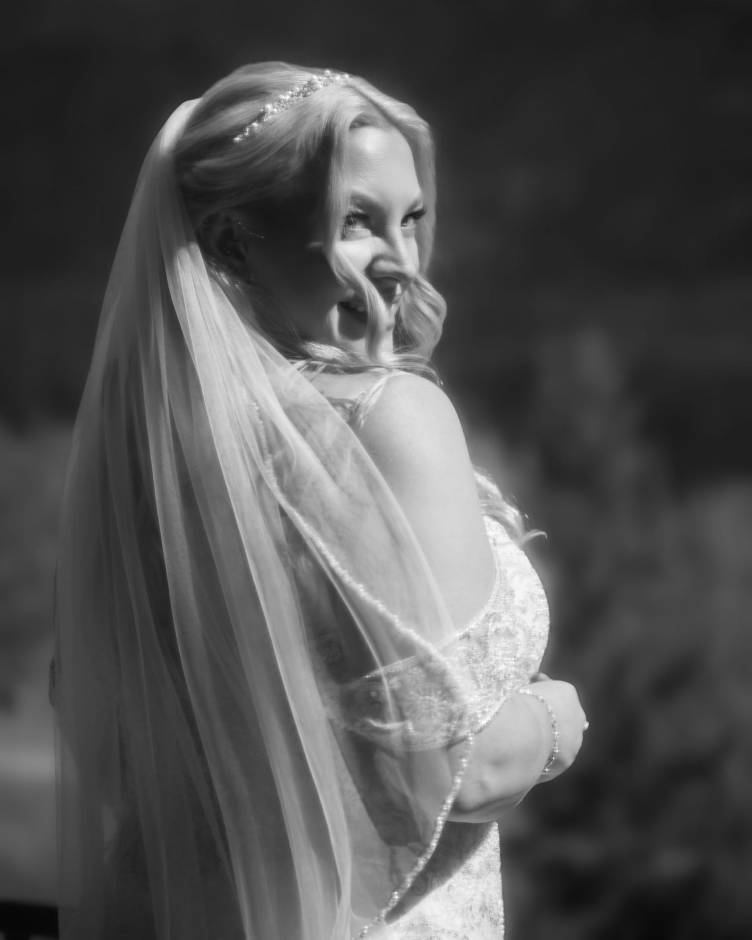Elegant bride in lace wedding gown and veil smiling over her shoulder in black and white photo.