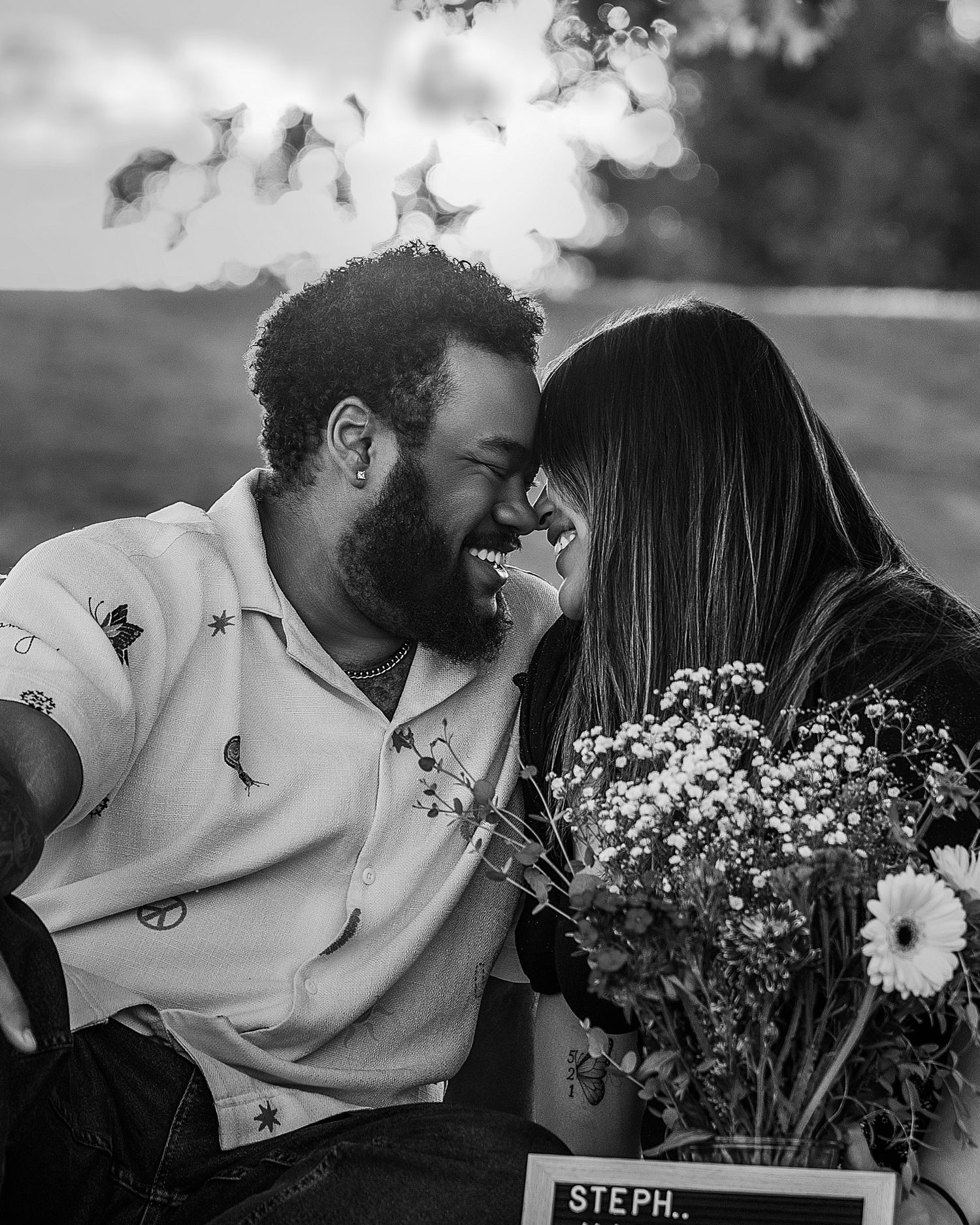 Black and white photo of joyful couple touching foreheads, woman holds bouquet with name sign visible.